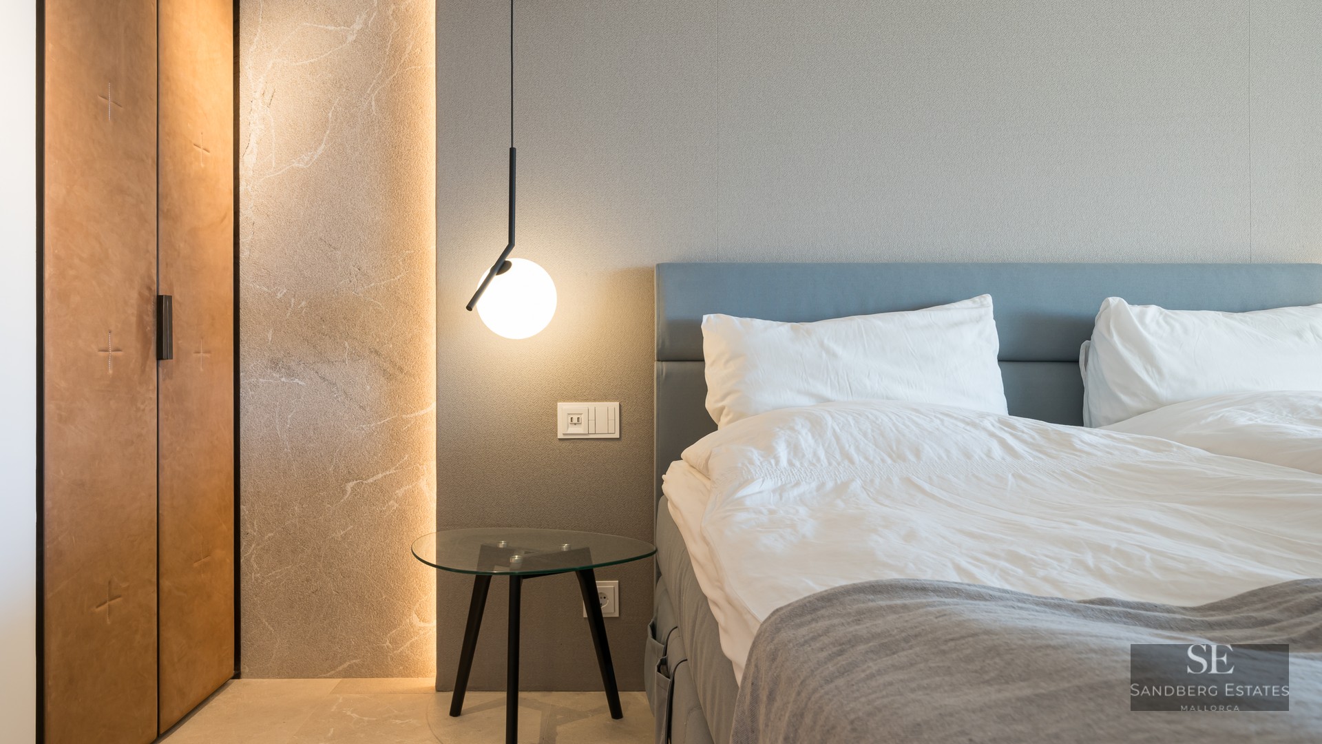 Luxury bedroom featuring a blue headboard, glass table, modern pendant light, and illuminated stone wall detail.