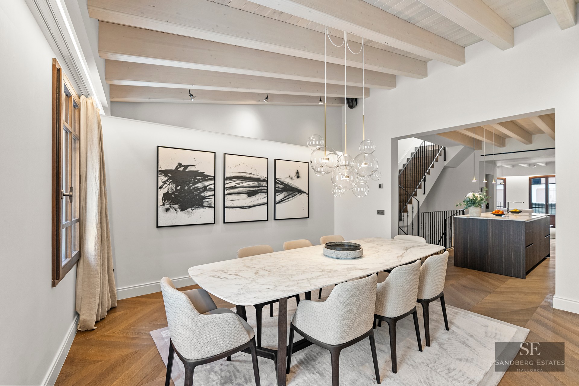 Modern dining room featuring a white marble table, designer chairs, bubble chandelier, and exposed wood ceiling beams.