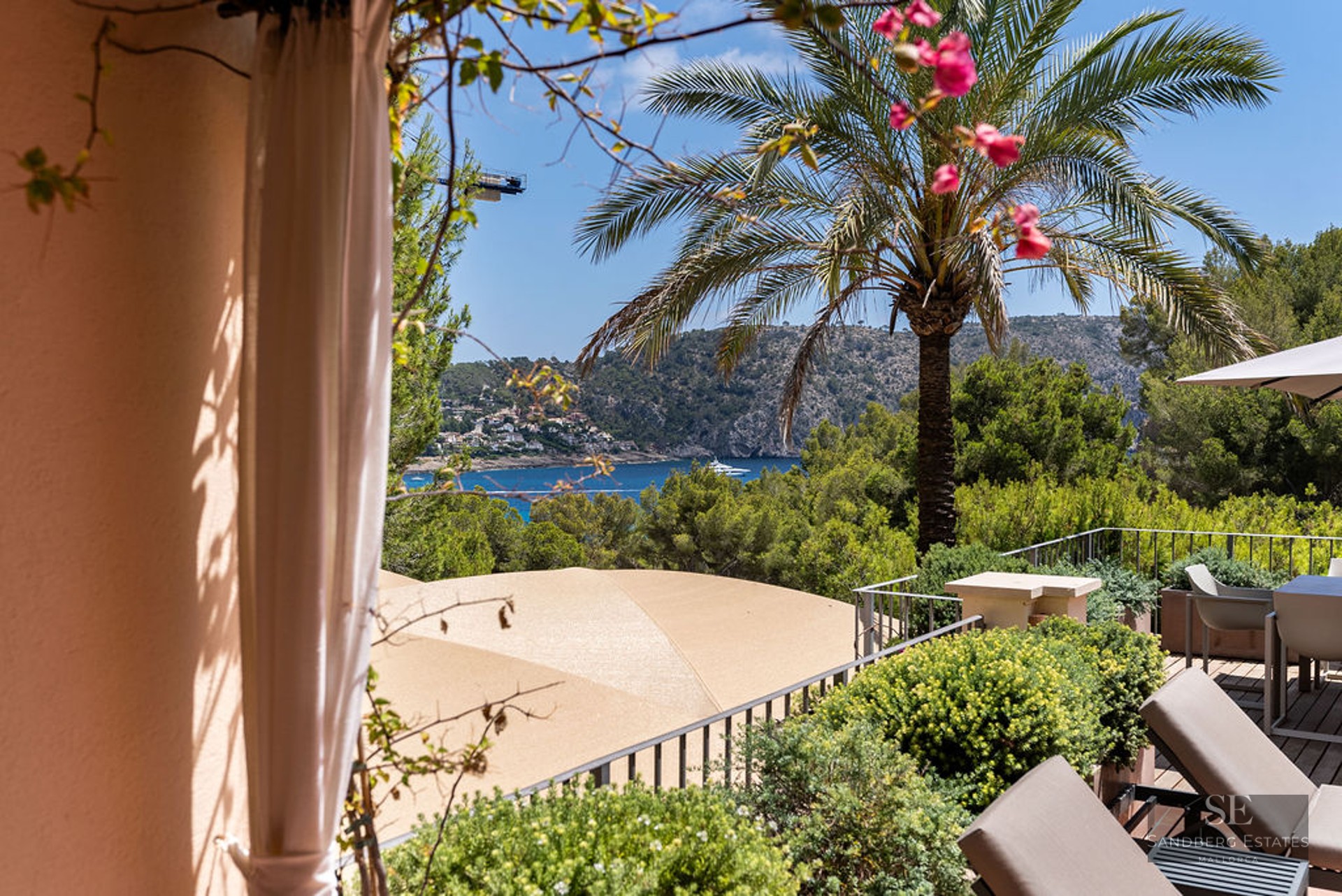 A luxury balcony terrace with lounge chairs and dining area overlooking a blue sea bay and green mountains.