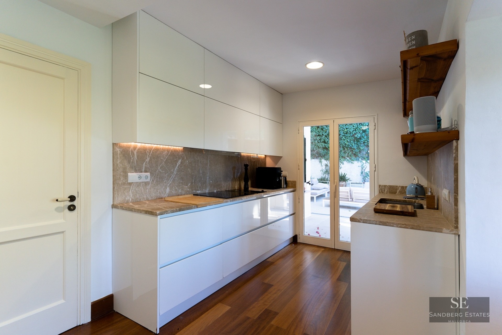 Modern white kitchen featuring marble countertops, wood flooring, and glass doors leading to an outdoor terrace.