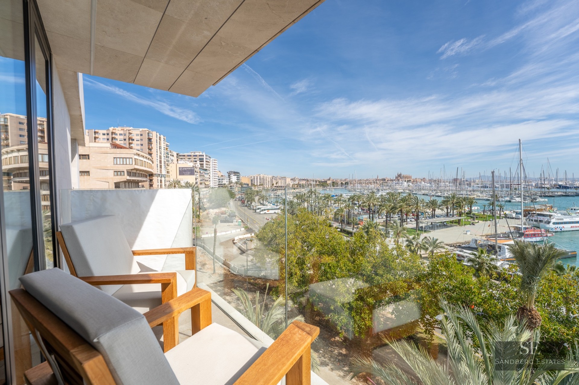 Modern balcony with wooden armchairs and glass railing overlooking a panoramic marina with yachts and city buildings.