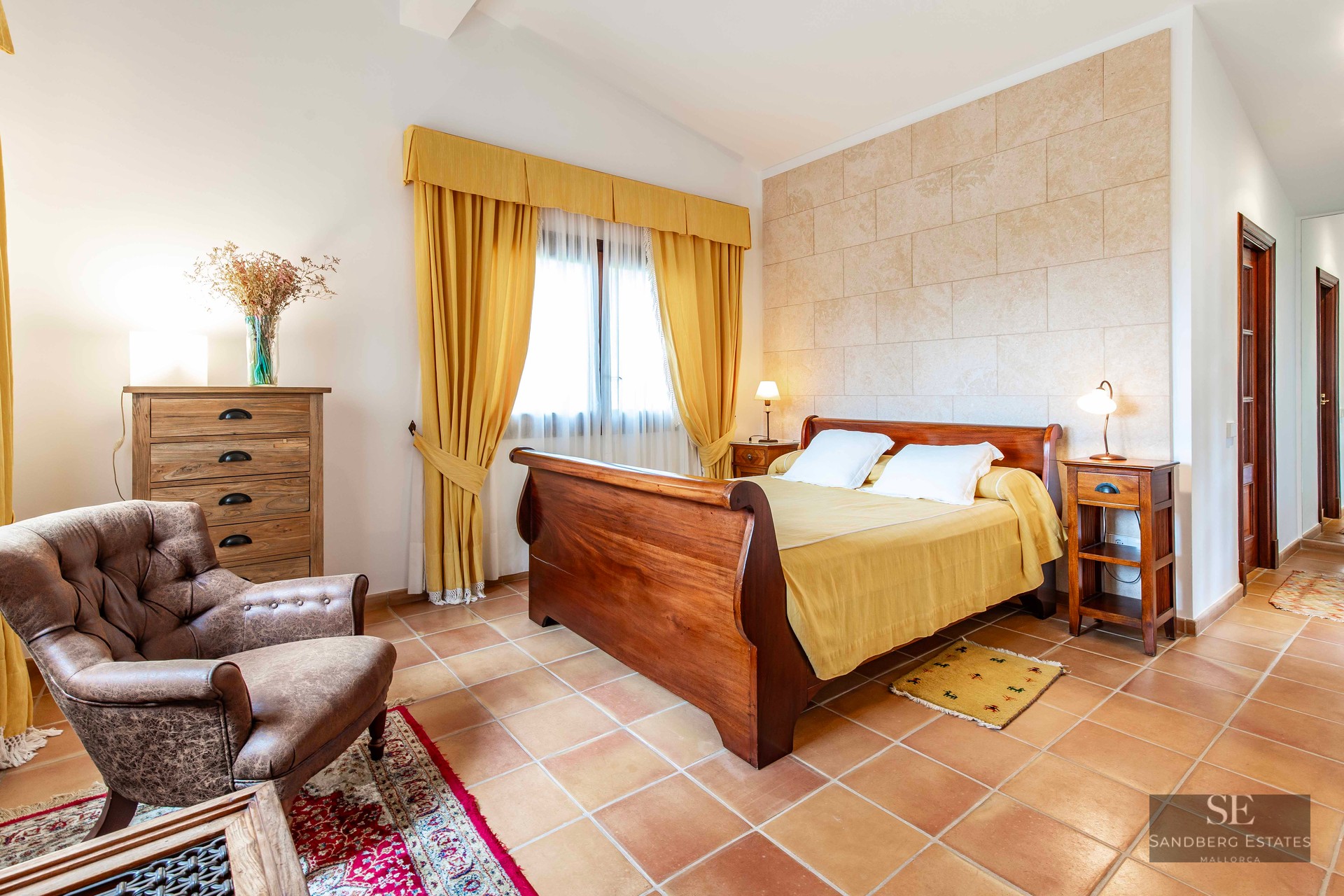 A rustic bedroom with a wooden sleigh bed, terracotta tiles, a leather armchair, and yellow curtains.
