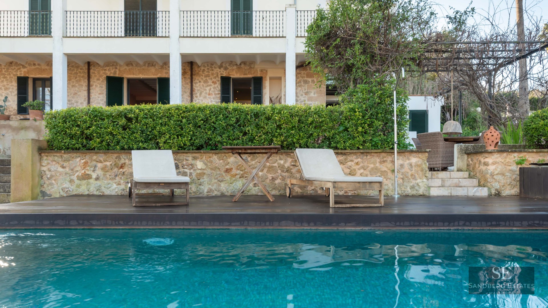 Turquoise swimming pool with wooden loungers in front of a stone Mediterranean house with green shutters.