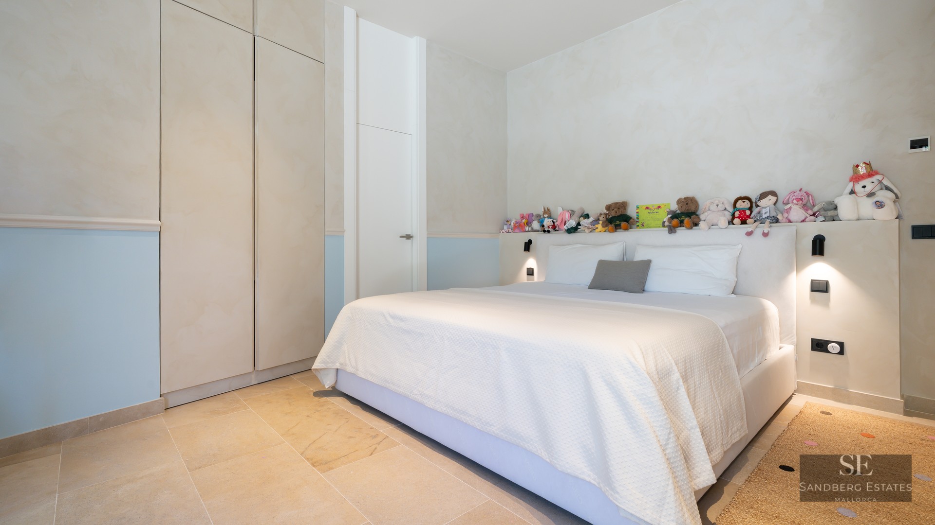 Bright bedroom featuring a large bed, stone floors, built-in wardrobes, and a row of stuffed animals on the headboard shelf.