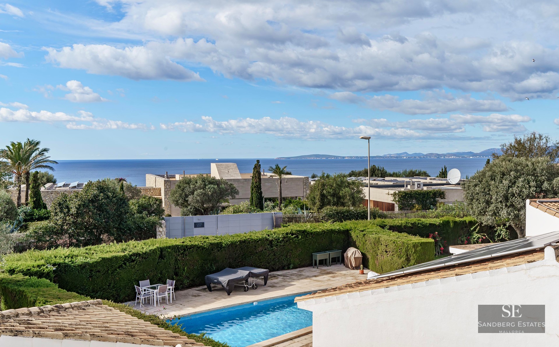 Elevated view of a blue swimming pool in a private garden with a panoramic ocean horizon under a cloudy sky.