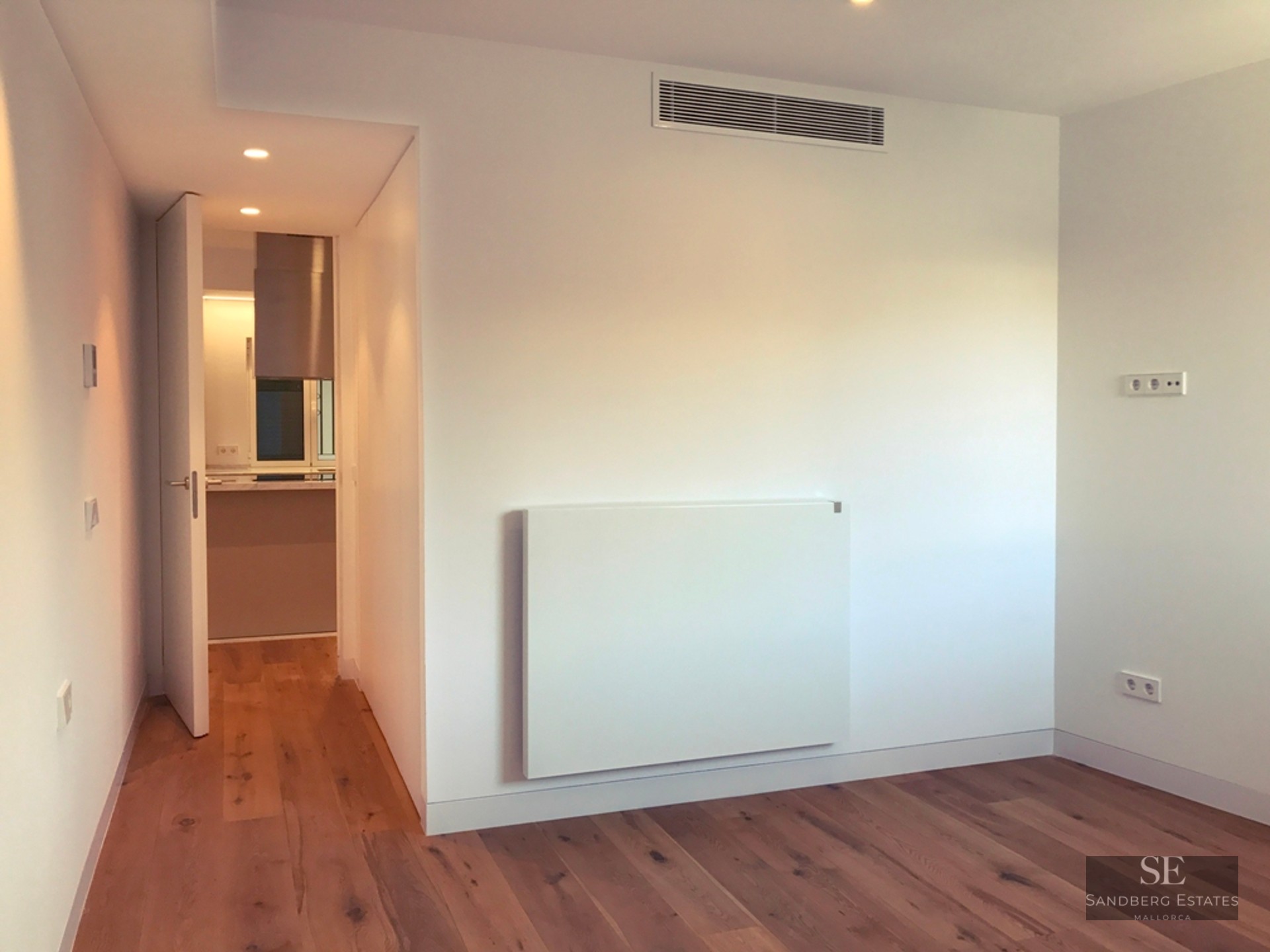 An empty room with white walls, light wood floors, and an open door leading to a kitchen area.
