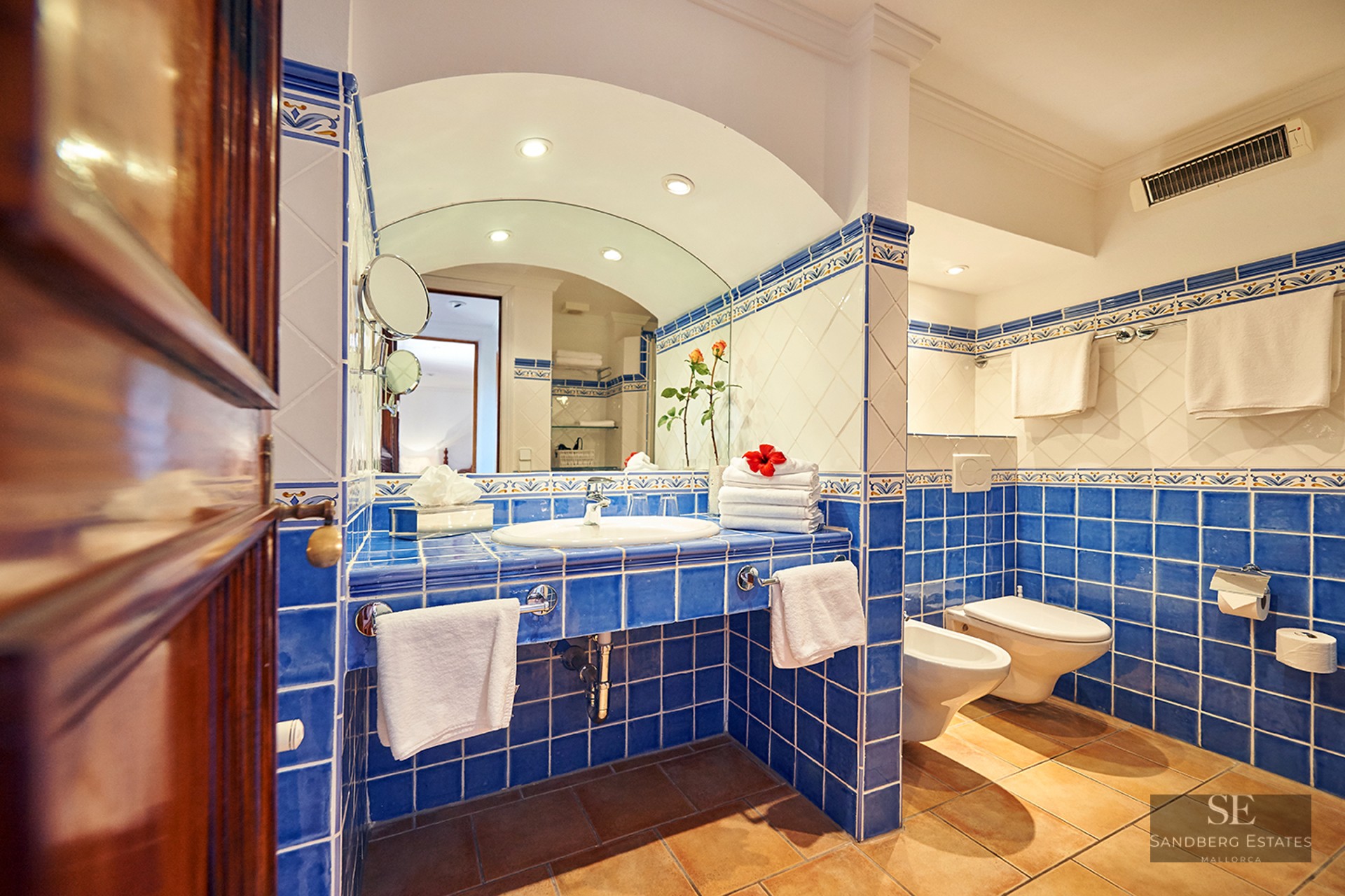 Bathroom with blue and white tiles, arched mirror, sink, bidet, and toilet over terracotta flooring.