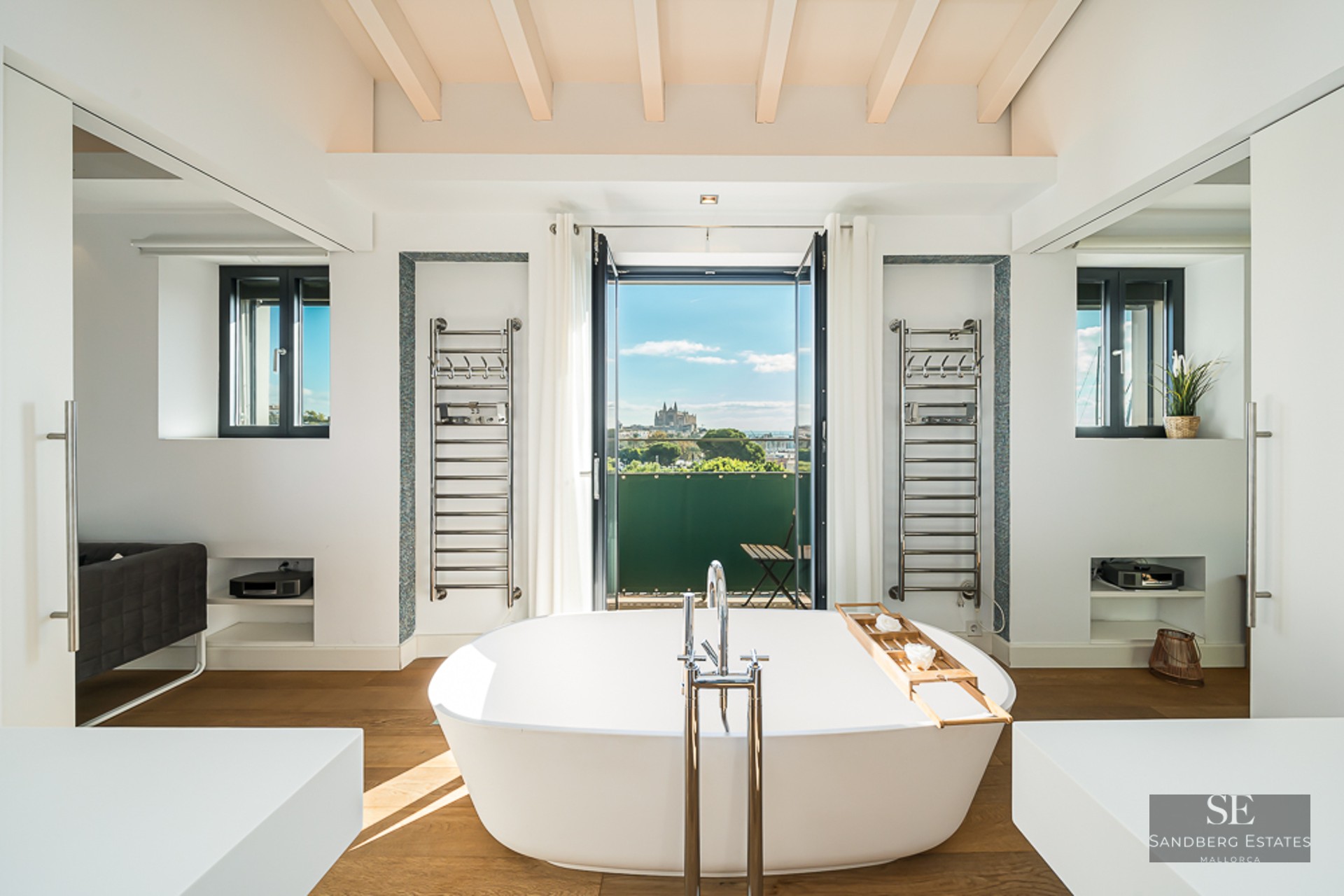 Freestanding white bathtub facing a balcony with city and cathedral views, featuring chrome towel rails.