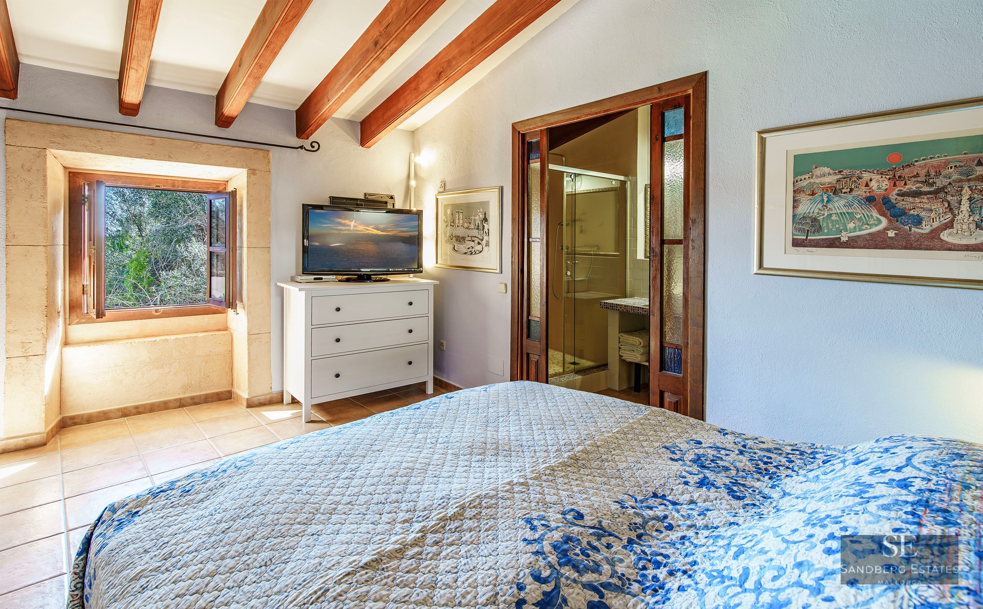 Bedroom with exposed wooden beams, a blue patterned bed, visible ensuite bathroom, and a stone-framed window.