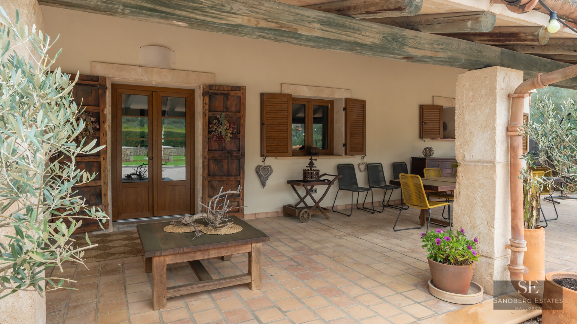 Covered porch featuring terracotta tiles, rustic wooden beams, dining furniture, and light stone walls.