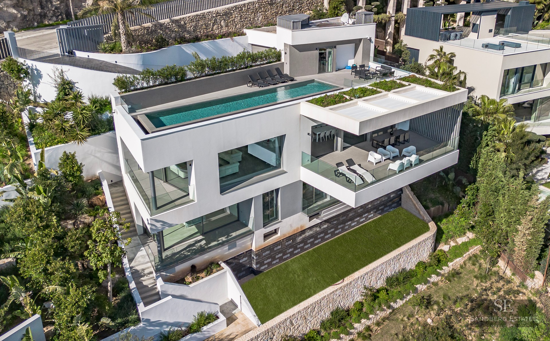 Aerial view of a white three-level modern villa with a rooftop infinity pool and glass-fronted balconies.