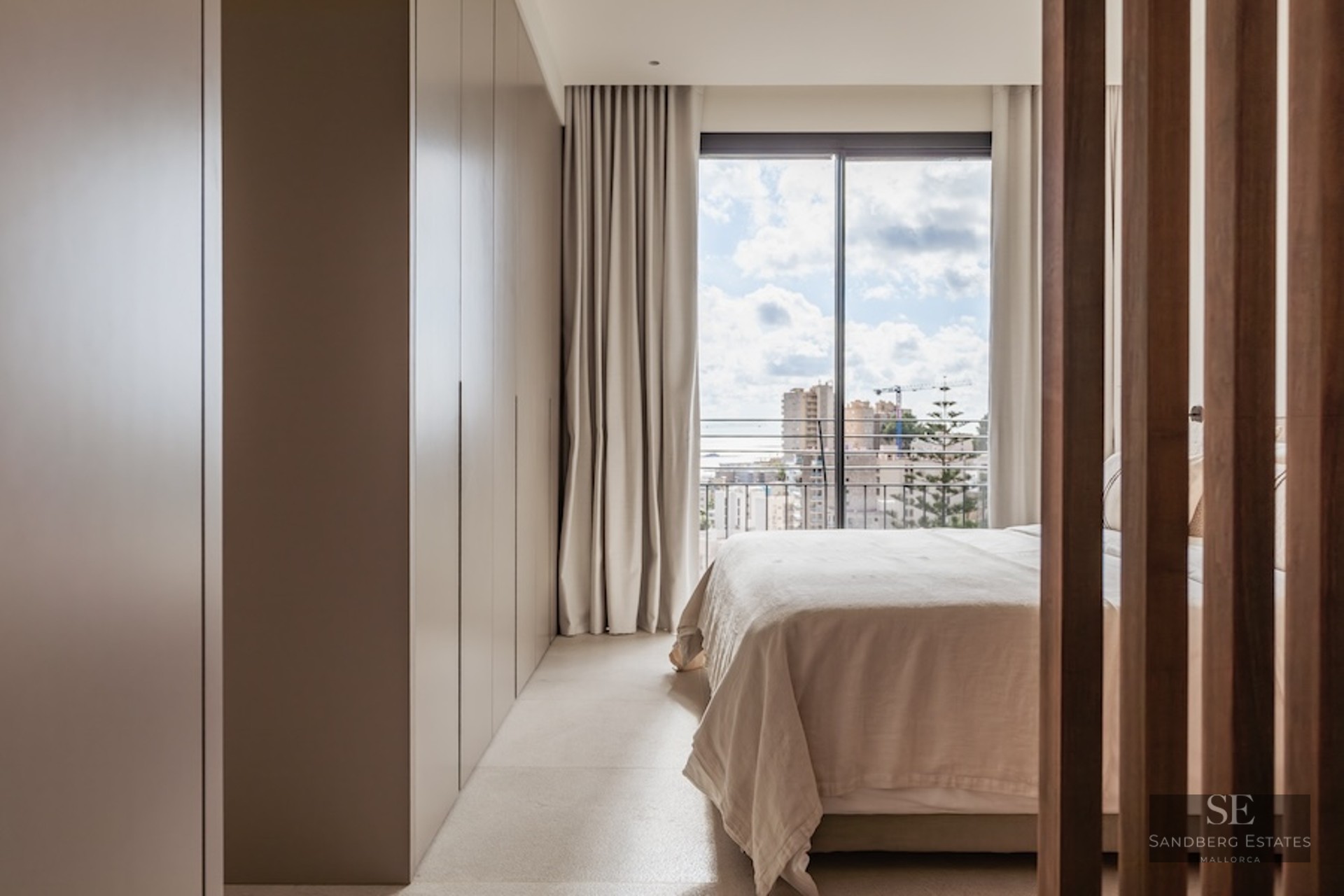 Bright bedroom with neutral tones, a large built-in wardrobe, and a floor-to-ceiling window overlooking a city.