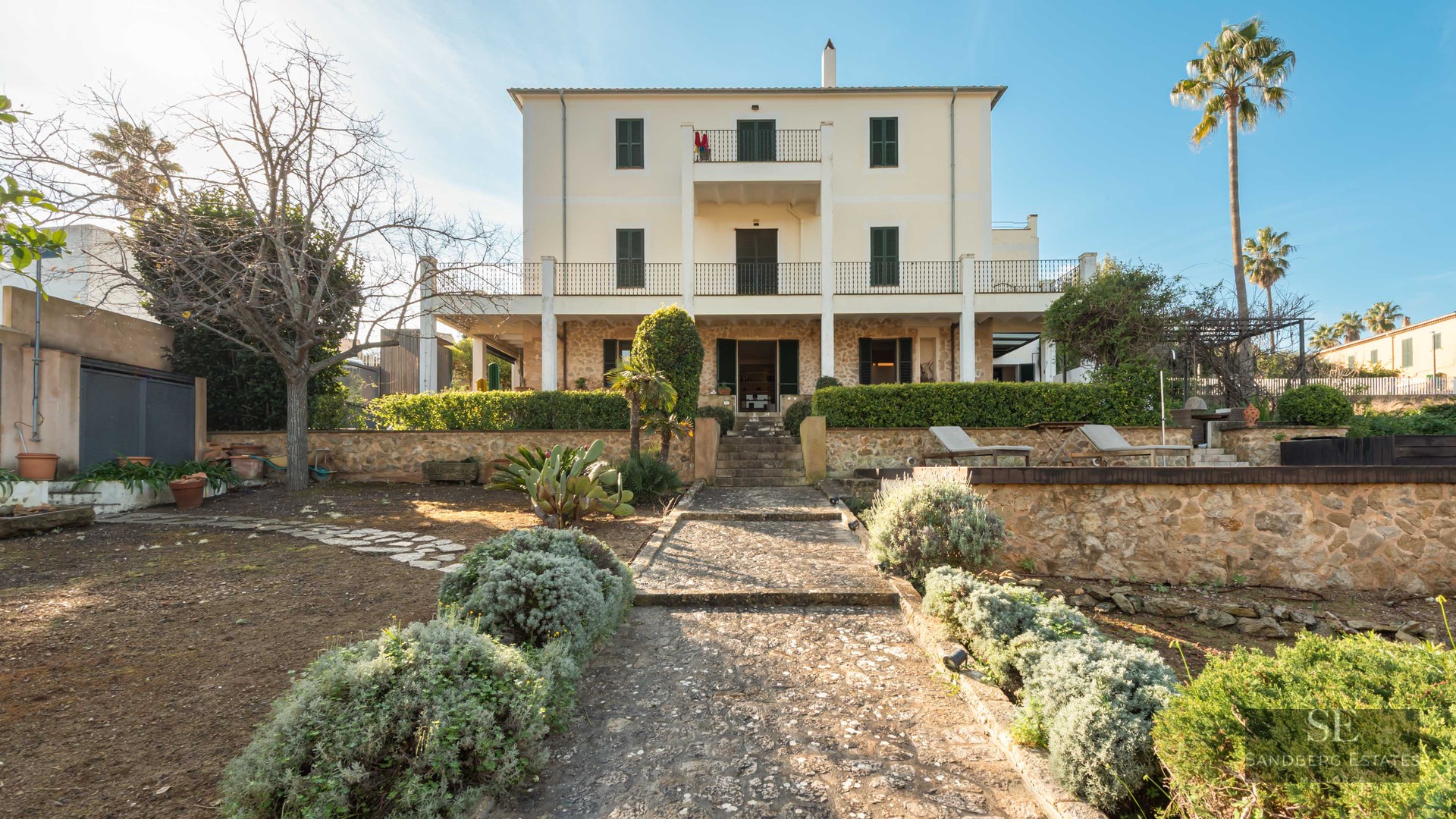 Rear view of a cream three-story villa with stone walls, green shutters, and a stone path through a garden.