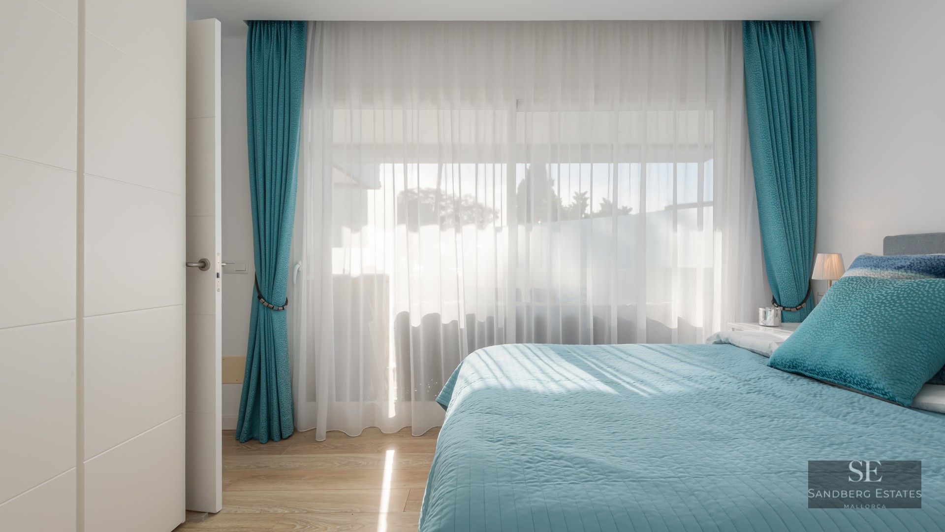 Chambre lumineuse avec lit turquoise, rideaux assortis et grandes fenêtres laissant entrer la lumière naturelle.