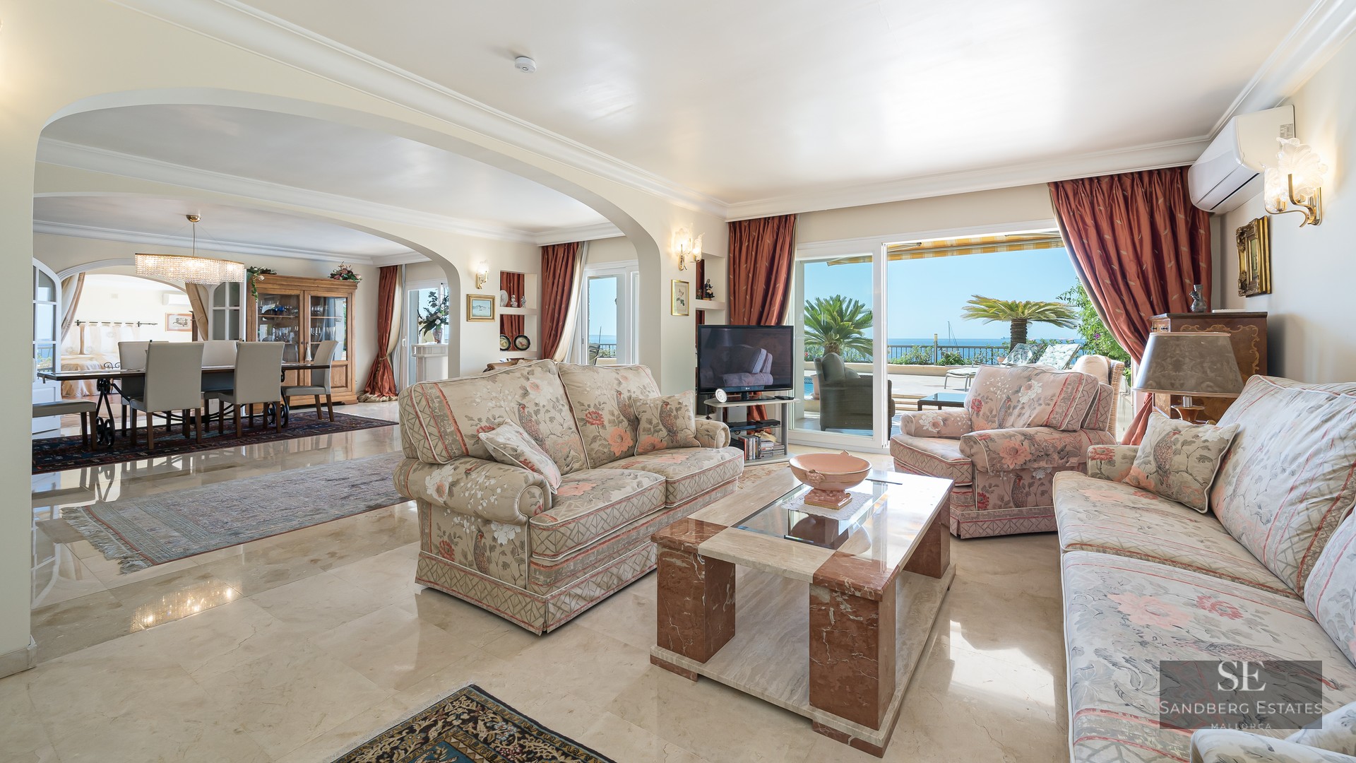 Bright living room with floral sofas, marble coffee table, and large glass doors opening to a terrace with sea view.