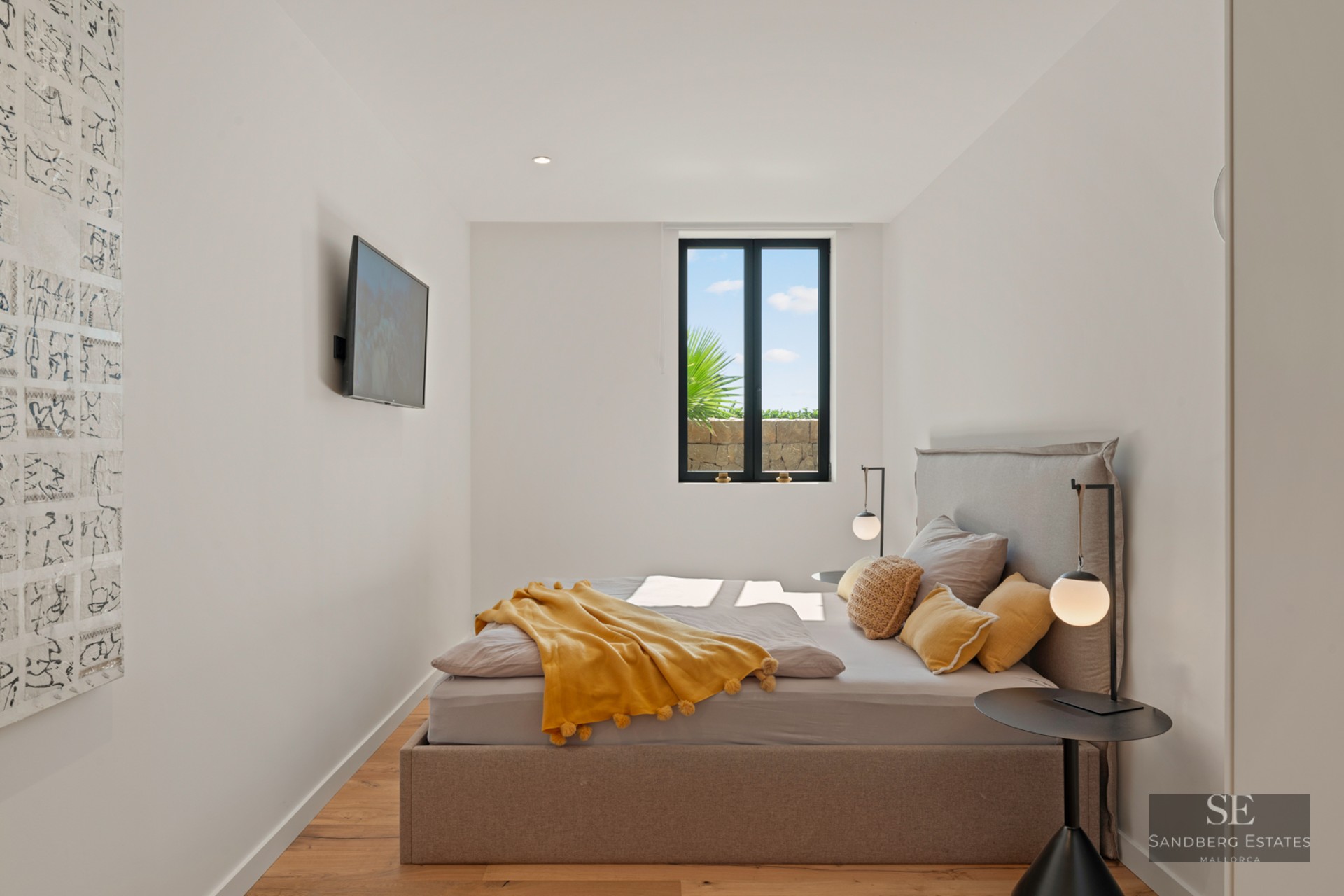 Bright bedroom featuring a taupe upholstered bed, yellow pom-pom blanket, minimalist lamps, and a scenic window view.