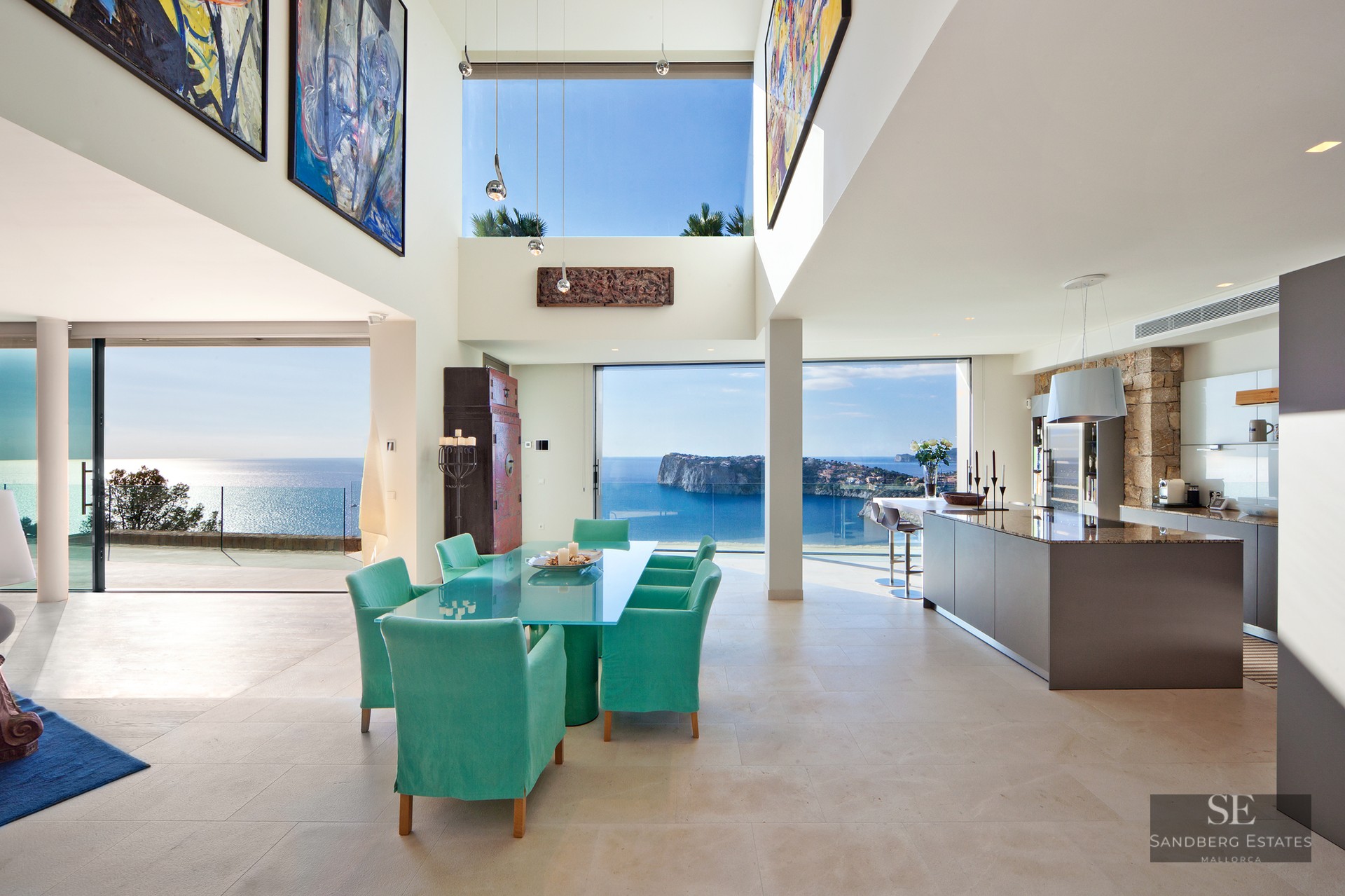 Modern open-plan dining and kitchen area with turquoise chairs and floor-to-ceiling windows overlooking the sea.