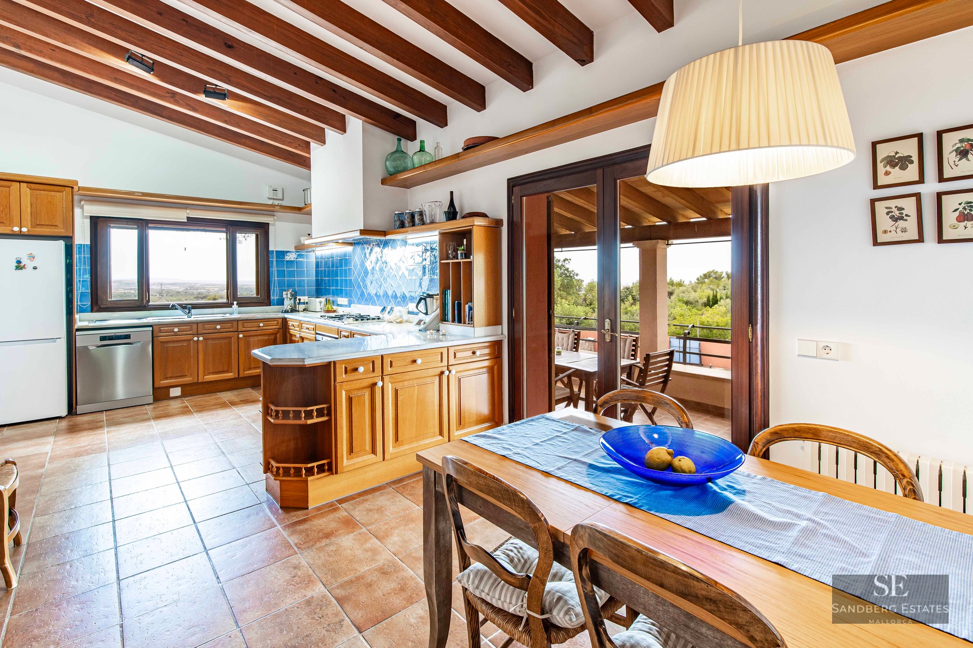 Kitchen with wood cabinets, blue tile backsplash, terracotta floor, and exposed wood beams with a dining table.