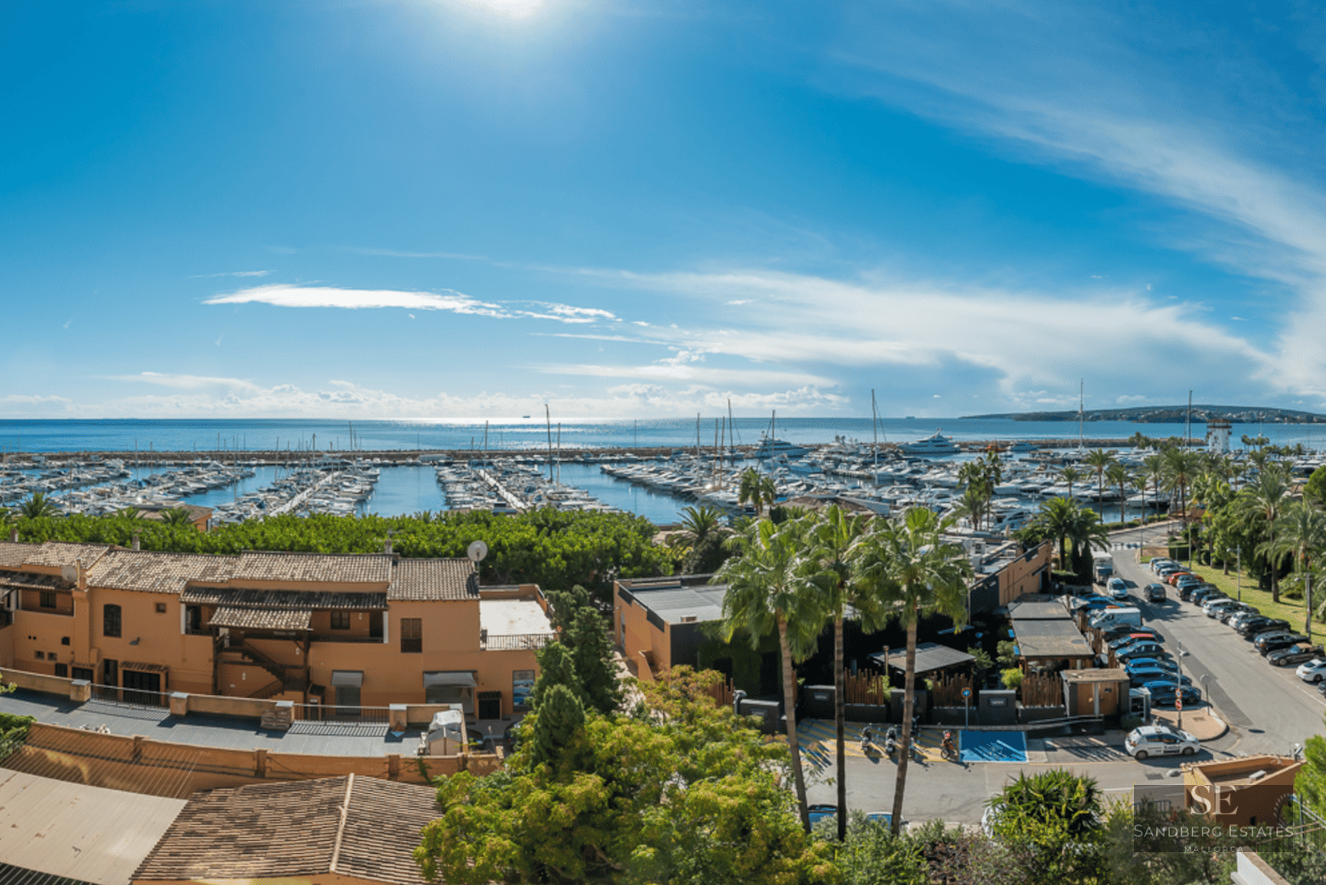 Elevated view of a Mediterranean port with yachts, terracotta buildings, and palm trees under a bright blue sky.