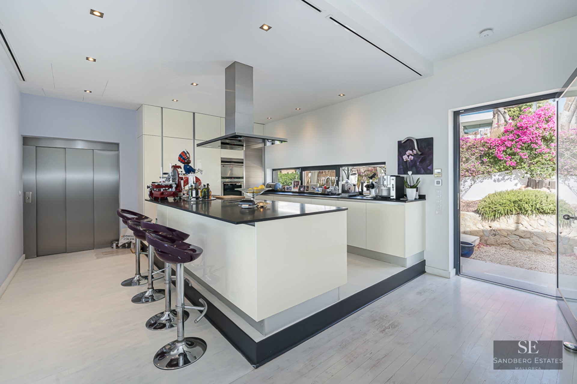 Contemporary white kitchen with a large central island, purple bar stools, stainless steel appliances, and home elevator.