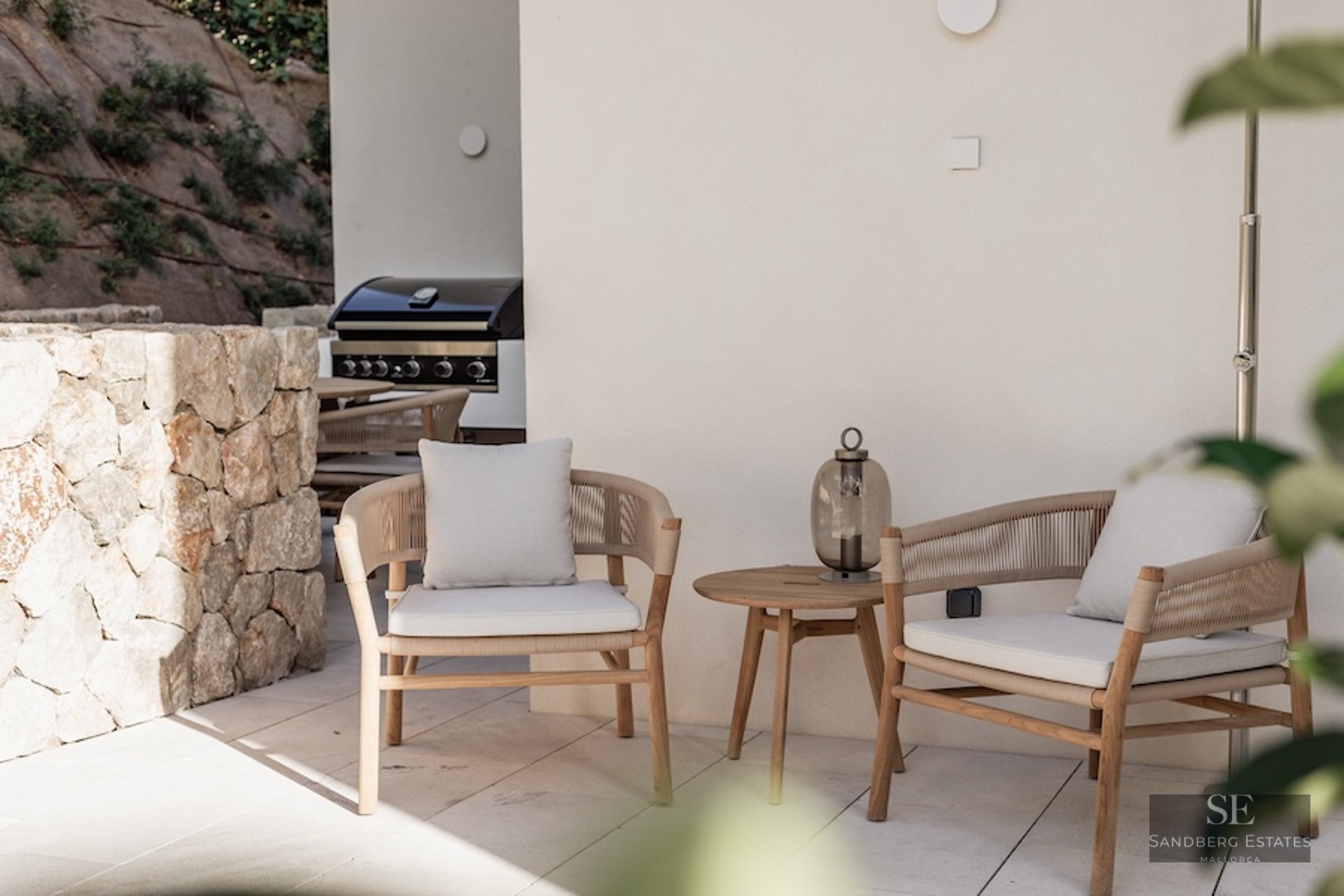 Two elegant modern armchairs with cushions next to a wooden side table on a stone terrace with a grill in the background.