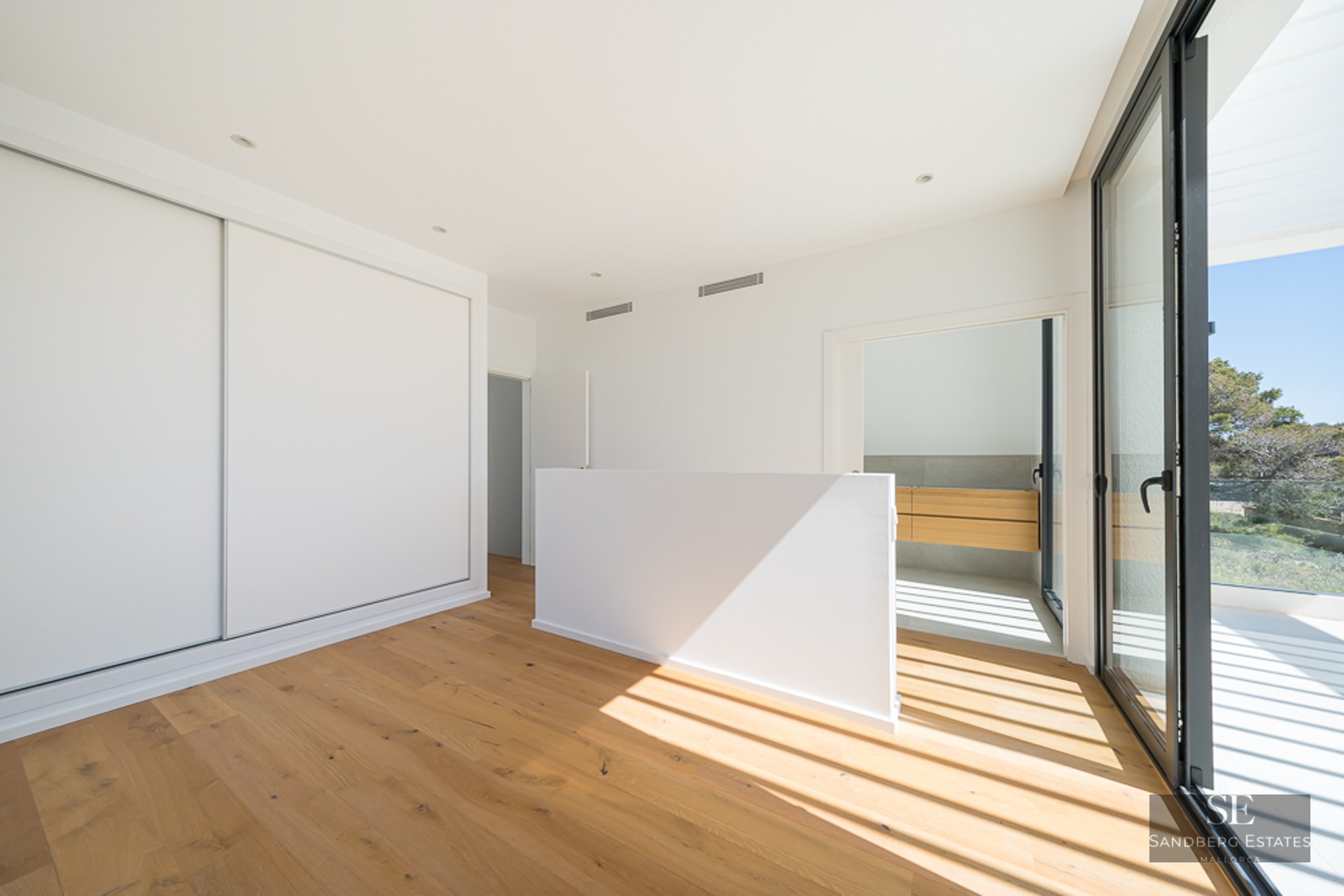 A bright, minimalist bedroom with oak floors, white sliding closets, and large glass doors opening to a terrace.