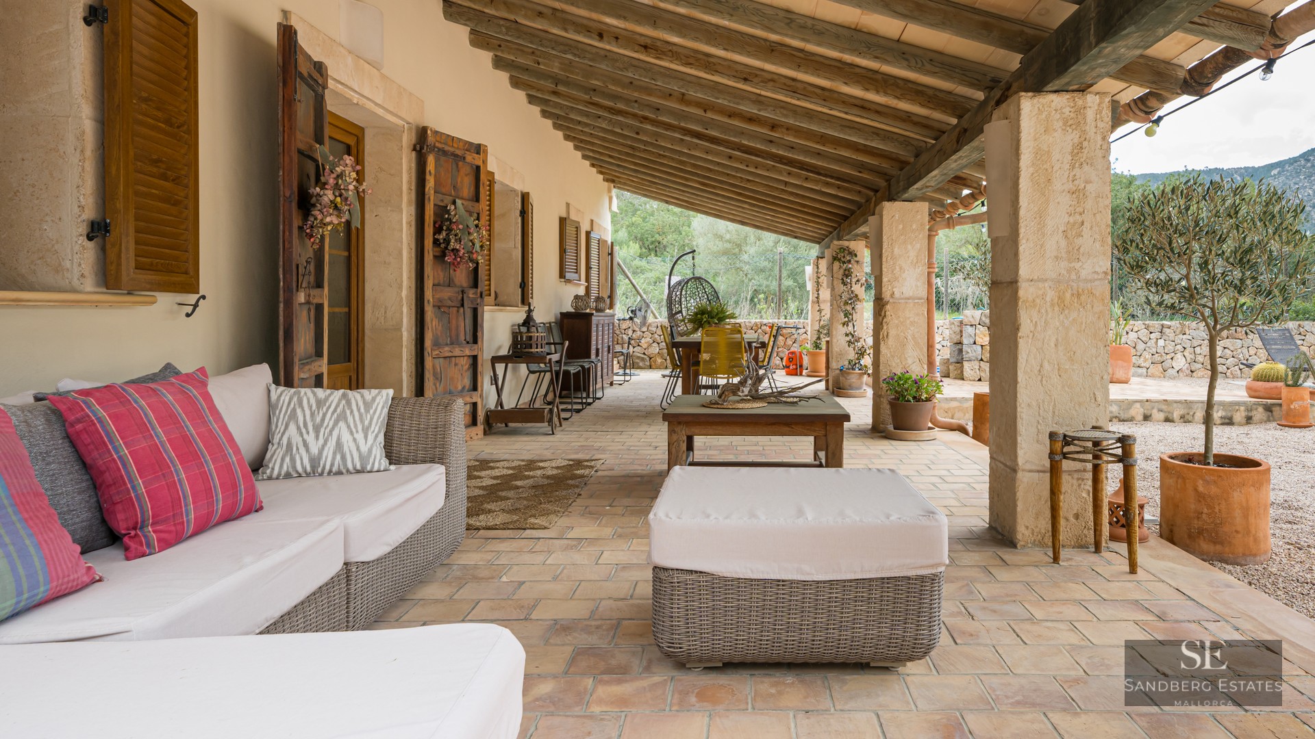 Shaded terrace featuring wicker sofas, exposed wooden beams, stone pillars, and terracotta tile flooring.