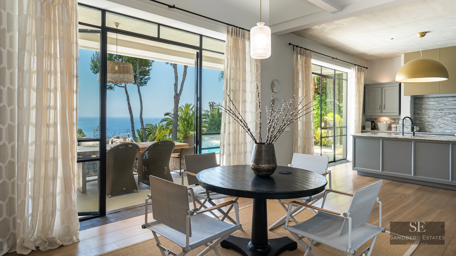 Modern dining room with a black round table and white chairs facing large glass doors with a view of the sea and terrace.