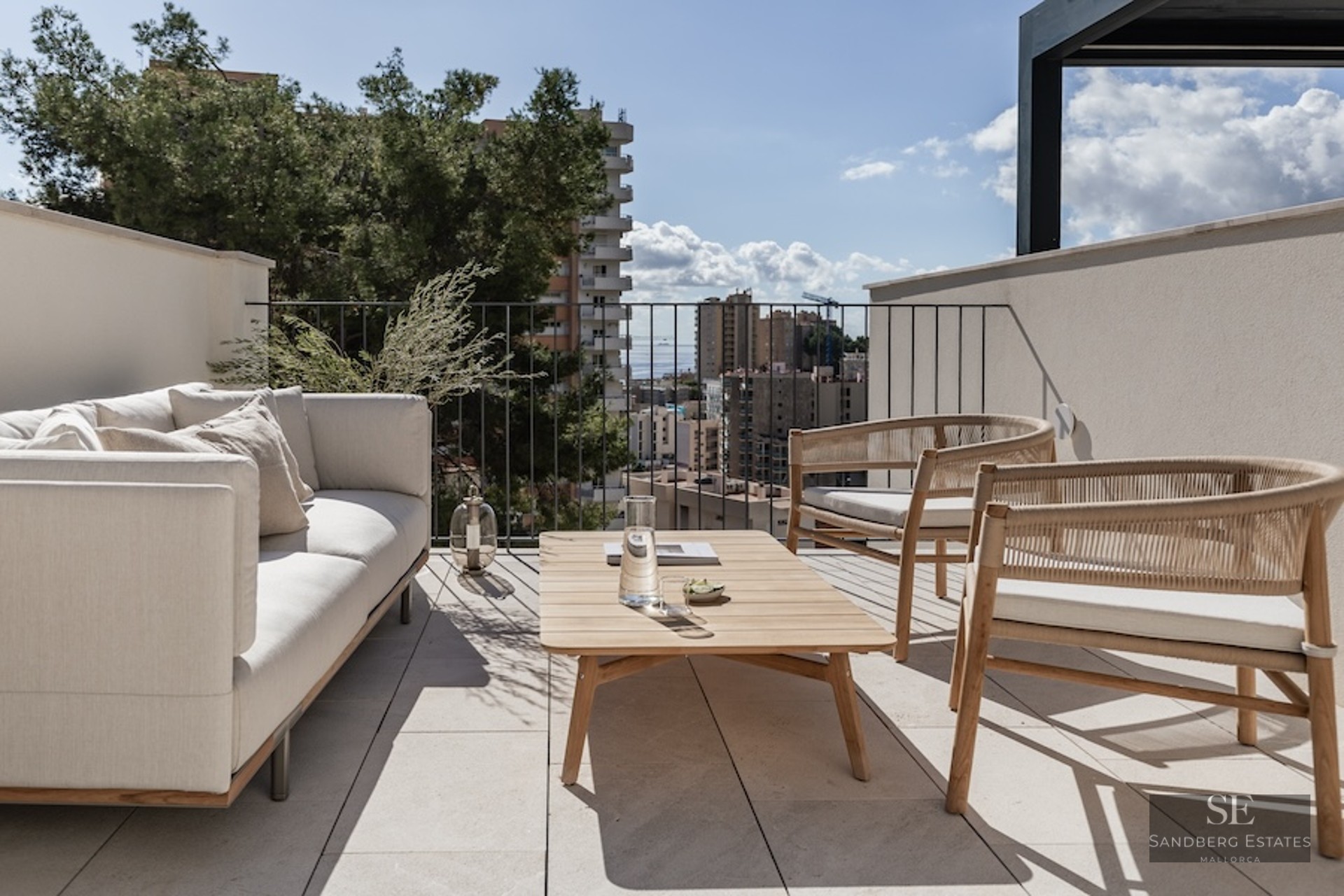 Luxury rooftop terrace with cream sofa, wooden armchairs, and a view of the city and sea under a bright sky.