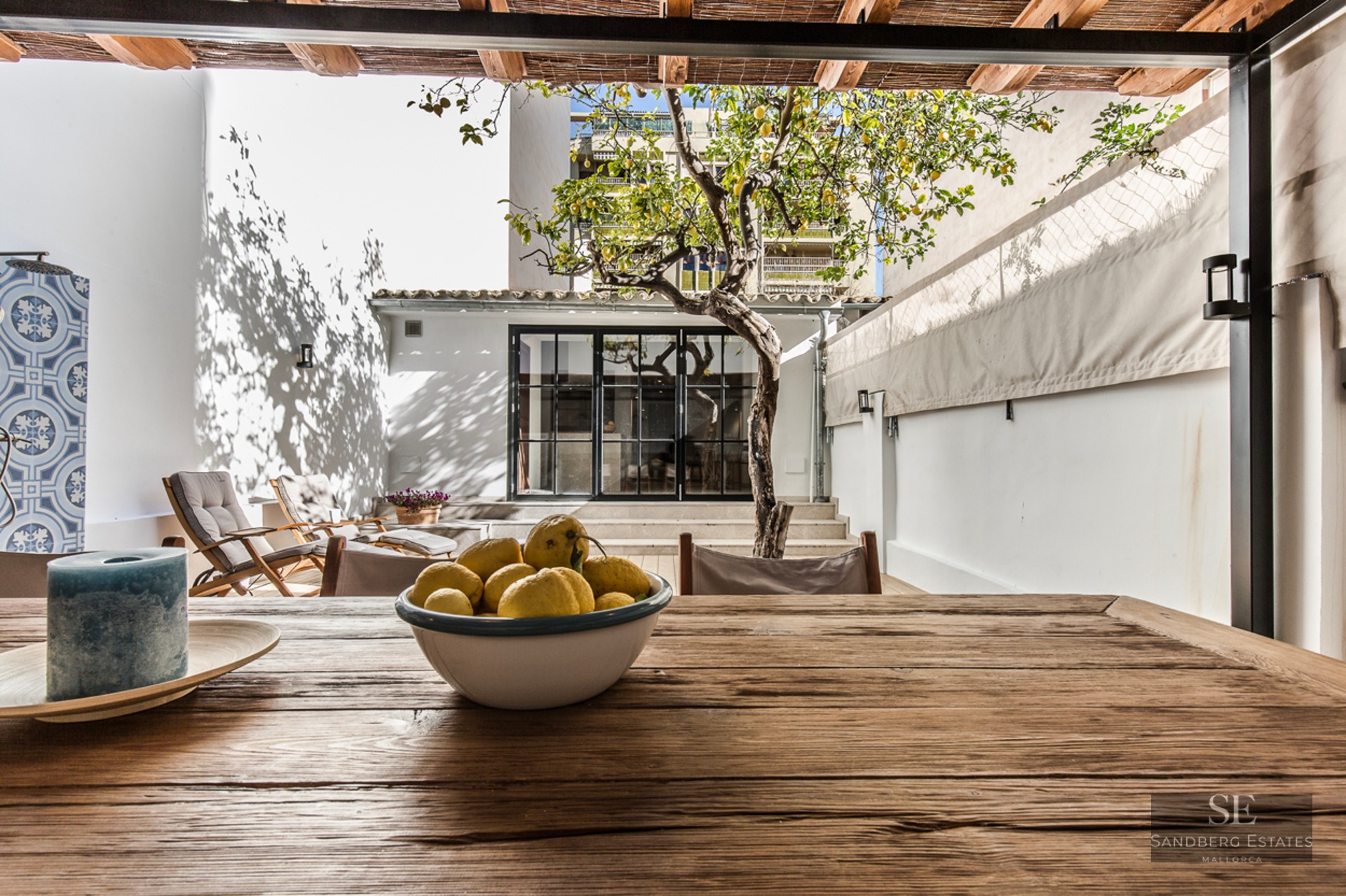 Sunny Mediterranean patio featuring a wooden dining table, a lemon tree, lounge chairs, and a white wall under a reed roof.