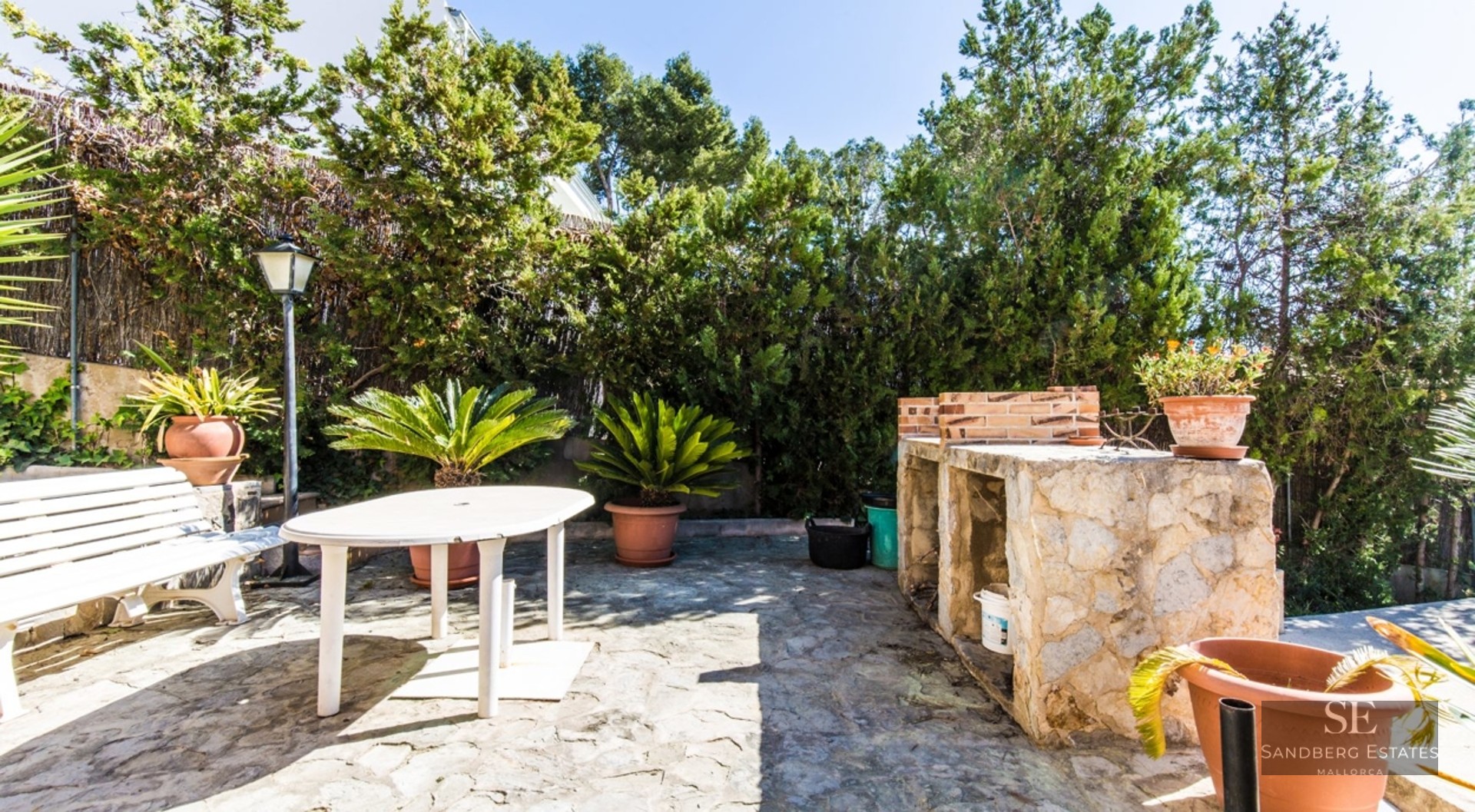 Paved patio with a stone barbecue, white outdoor table and bench, surrounded by lush green hedges and potted plants.