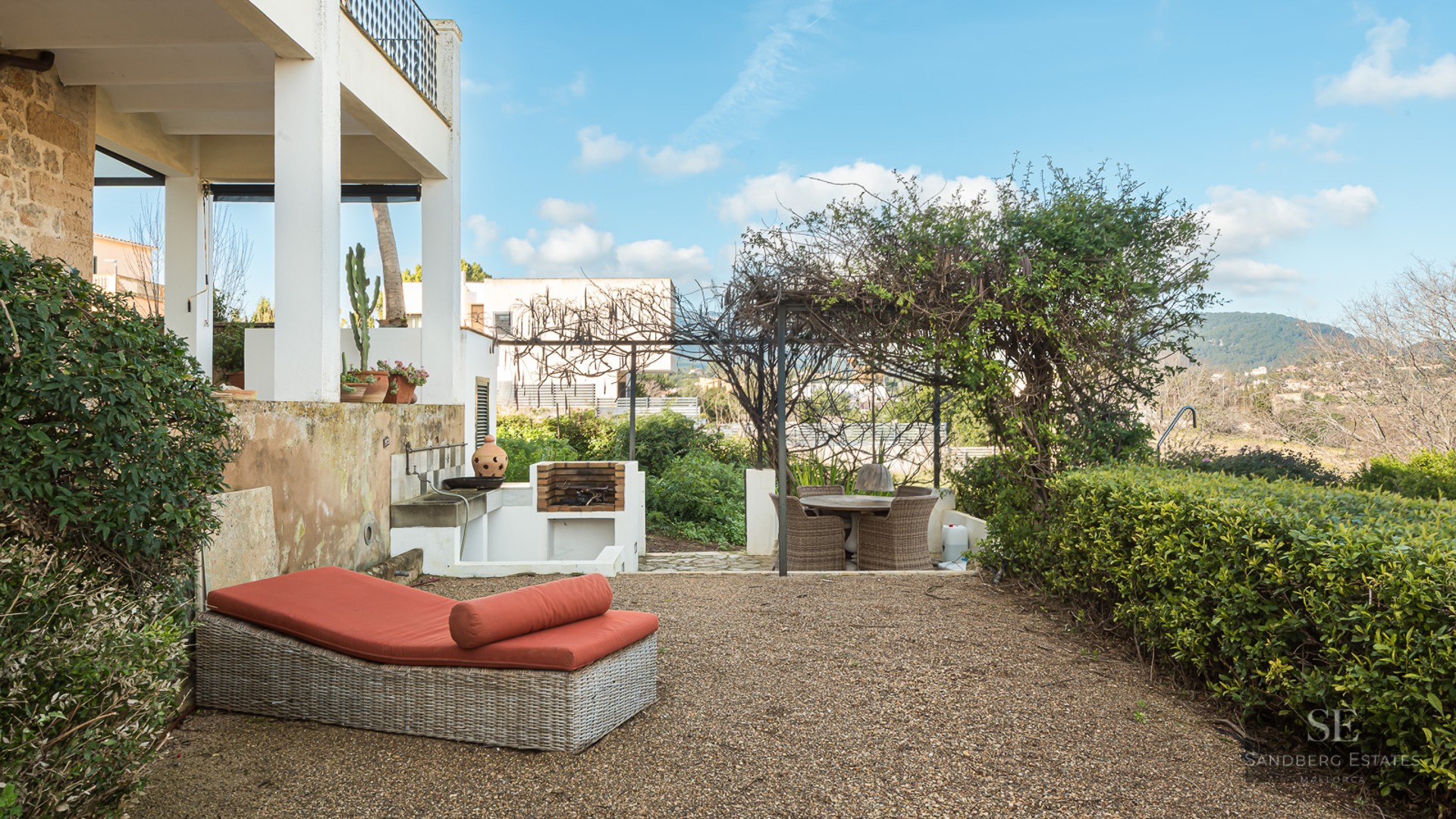 Outdoor terrace featuring a wicker sunbed with orange cushions, a built-in stone grill, and a pergola-shaded dining area.