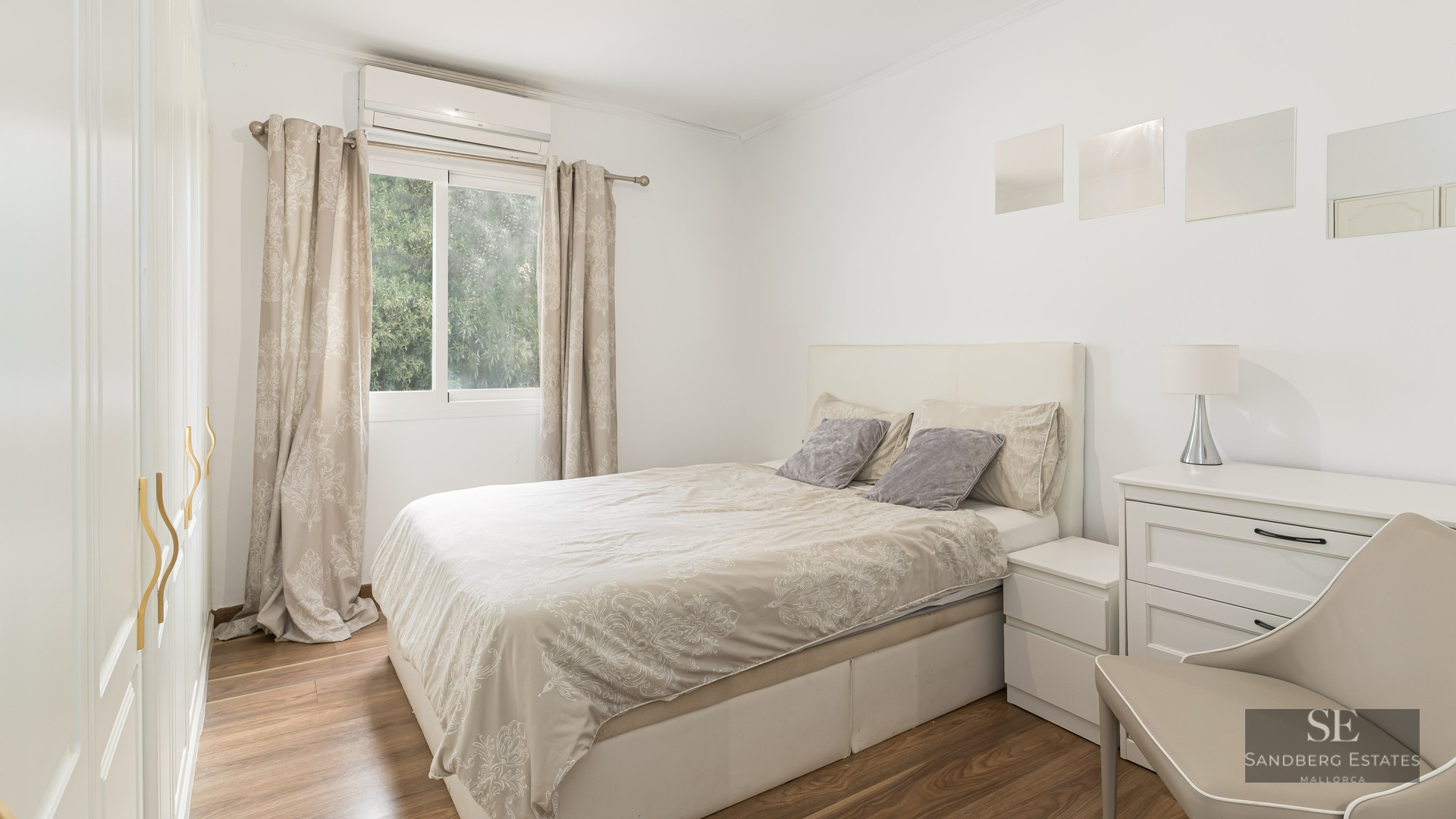 Chambre lumineuse avec lit double, meubles blancs, parquet et fenêtre donnant sur des arbres.