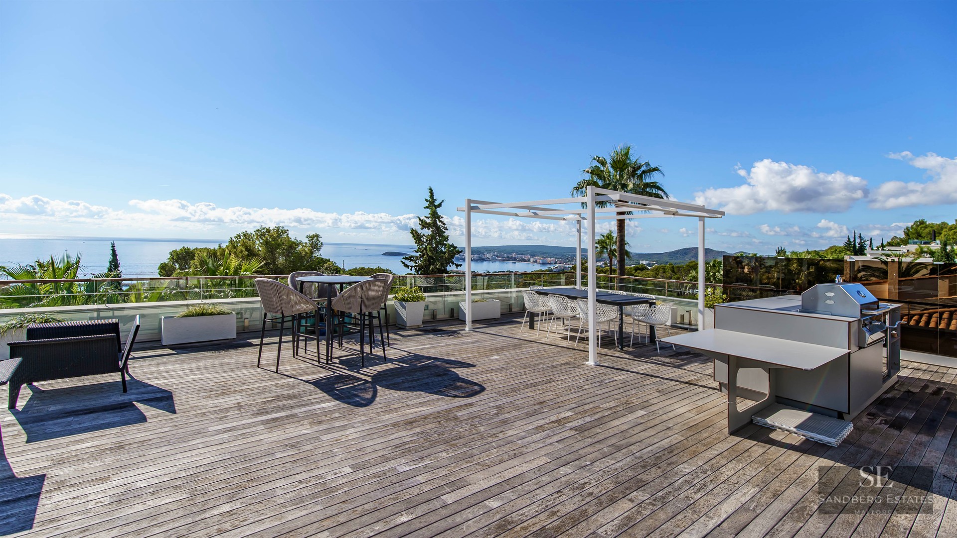 Grande terrasse en bois avec cuisine d'été, coin repas et garde-corps en verre surplombant l'océan.