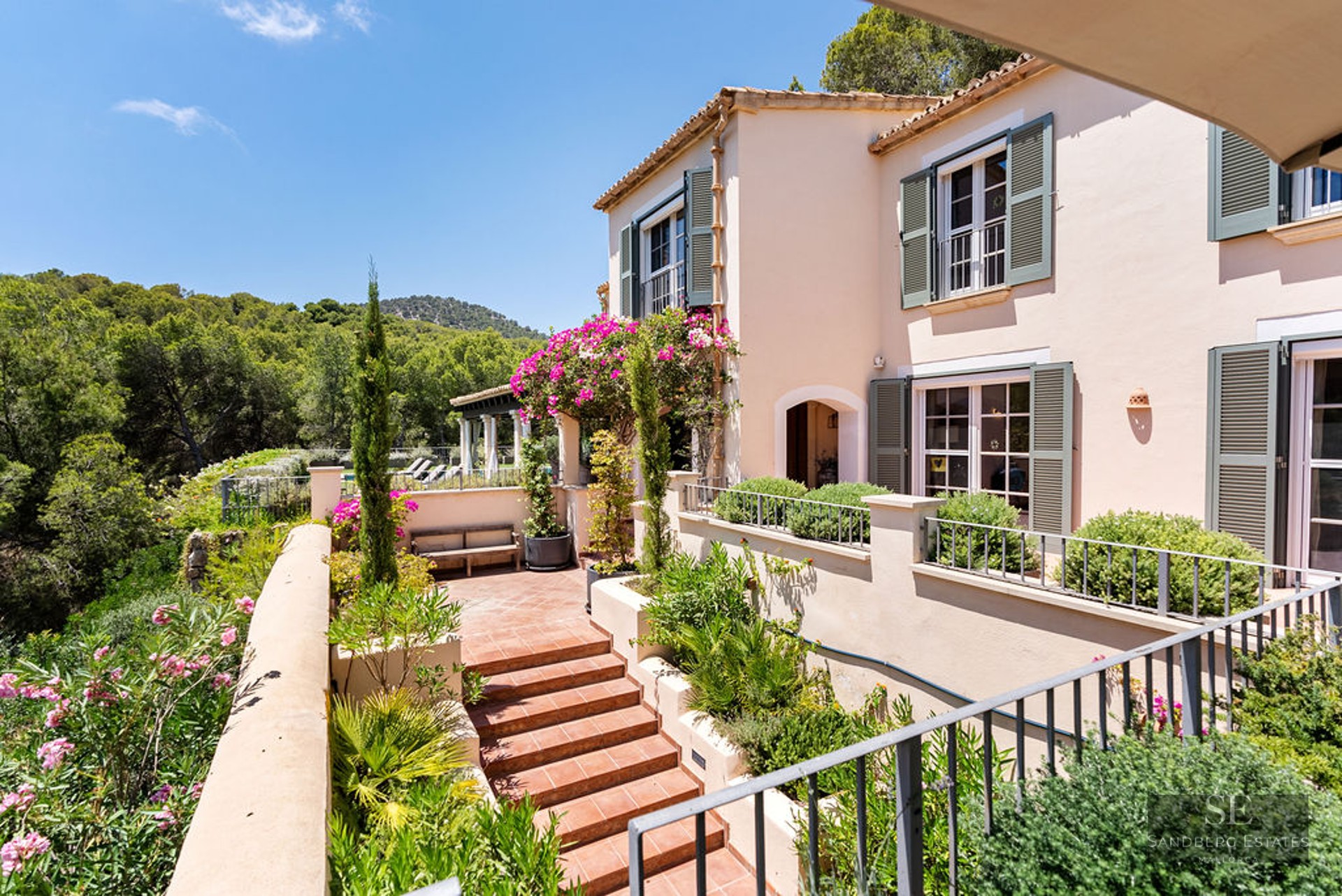 Sunny peach-colored villa with green shutters, terracotta stairs, and lush garden overlooking a forested valley.