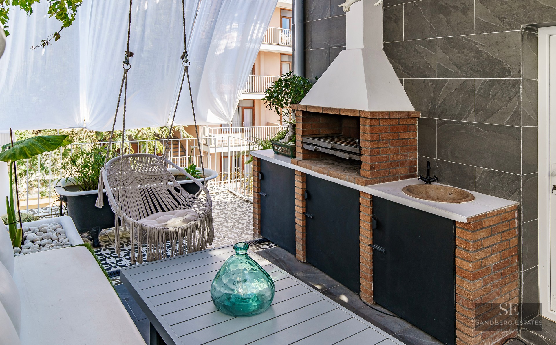 A modern terrace featuring a brick barbecue, a hanging macrame chair, a dining table, and decorative tiling.