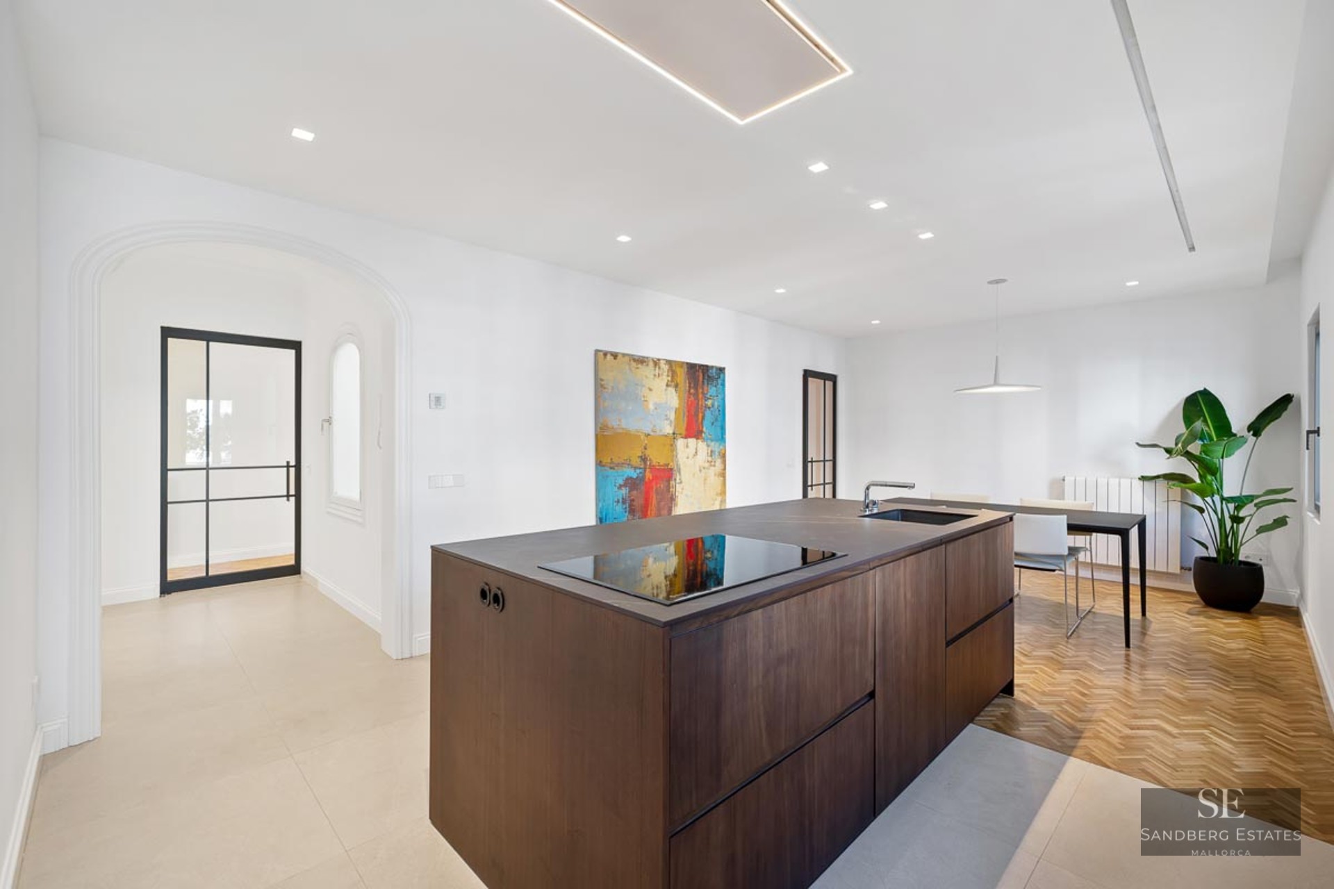 Luxury kitchen featuring a large dark wood island, herringbone flooring, and a modern dining area with a tall plant.