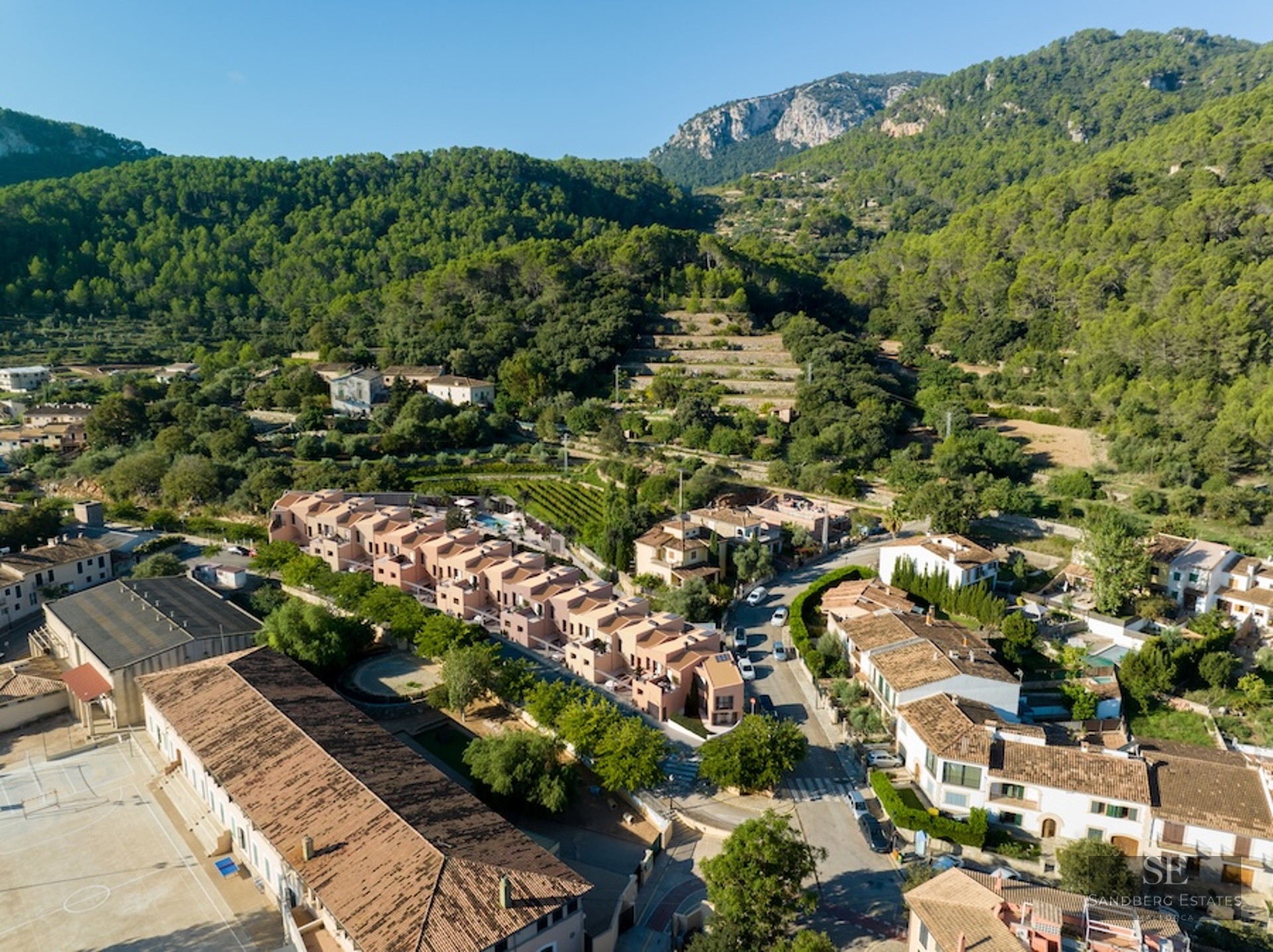 Aerial drone shot showing modern terracotta houses in a Mediterranean village surrounded by forested mountains.