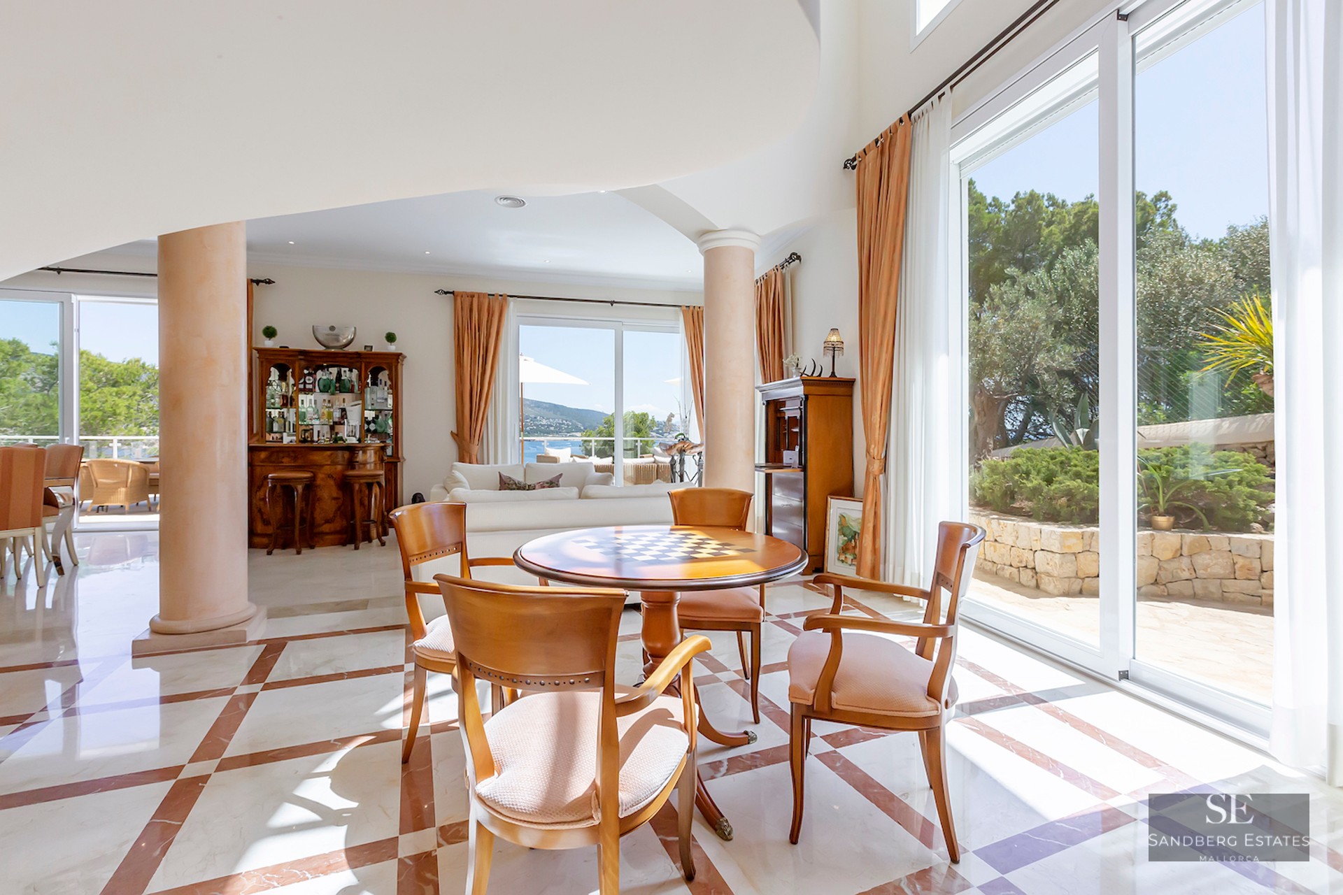 Spacious living area with marble floors, wooden table, pillars, and large windows overlooking a garden and sea.
