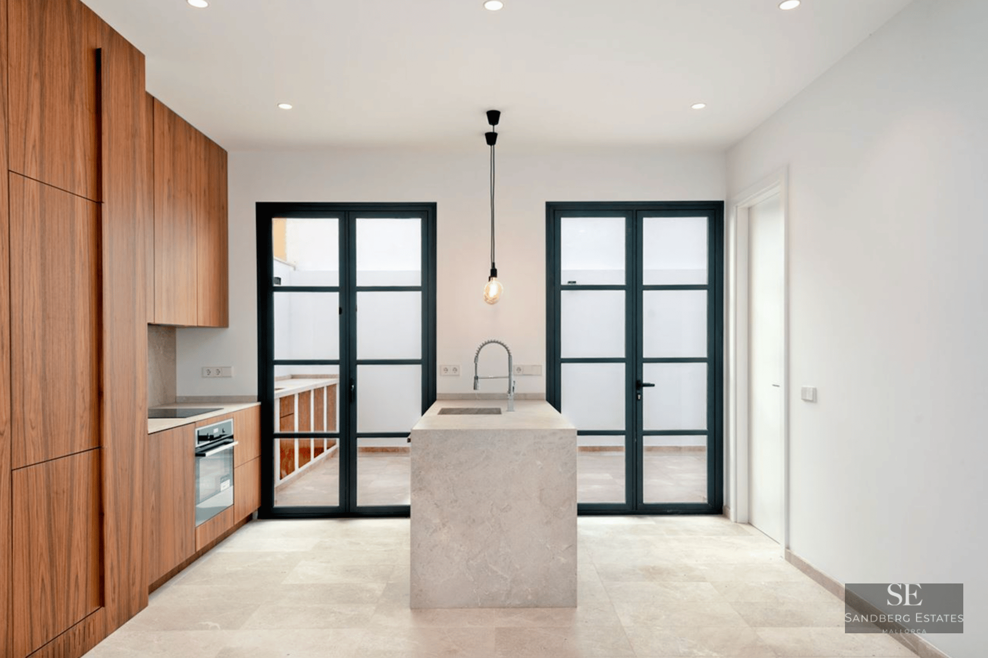 Minimalist kitchen featuring a central beige stone island, walnut wood cabinetry, and large black-framed glass doors.