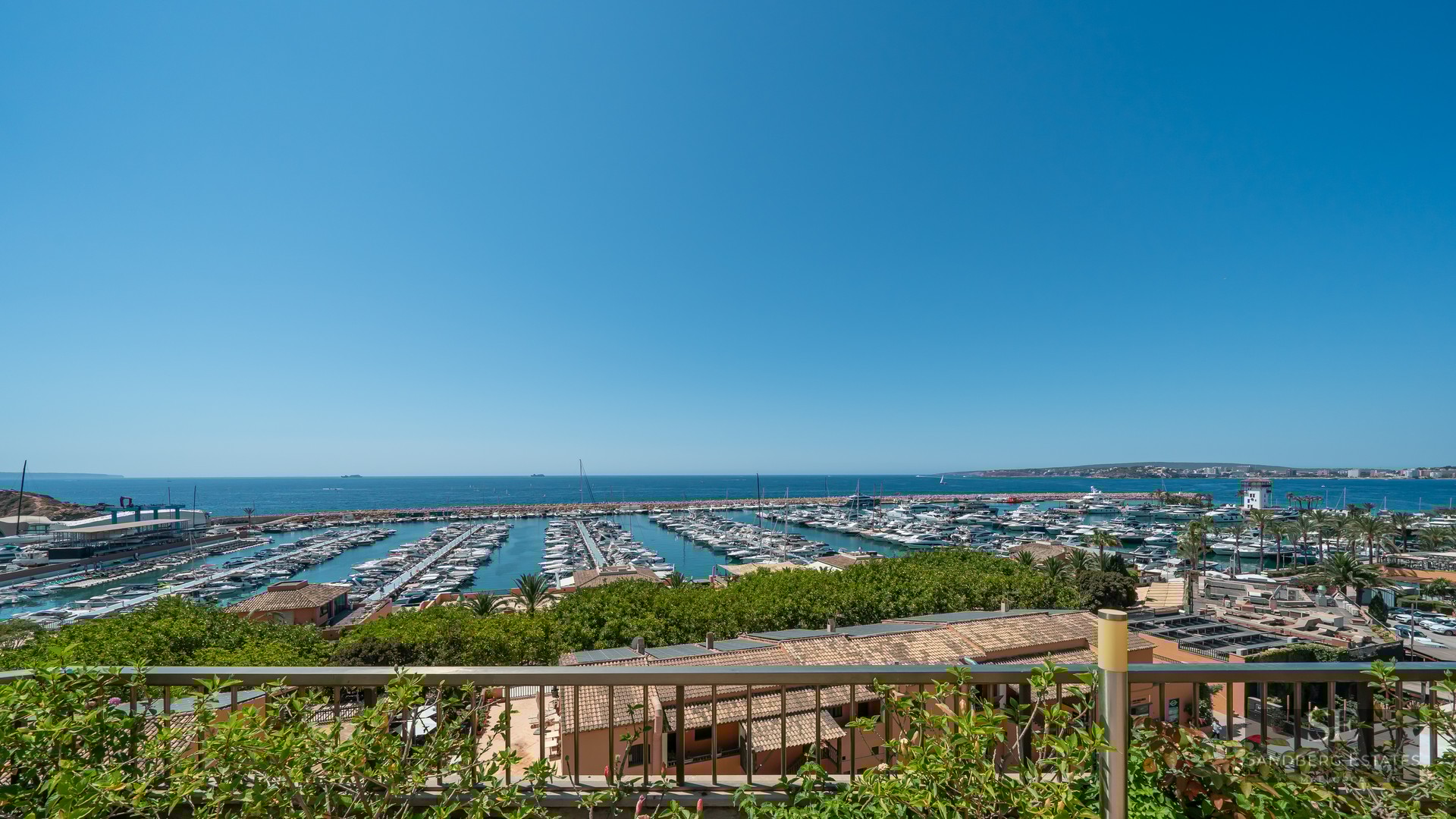 High-angle view of a bustling marina with white yachts under a clear blue sky, seen from a terrace with greenery.