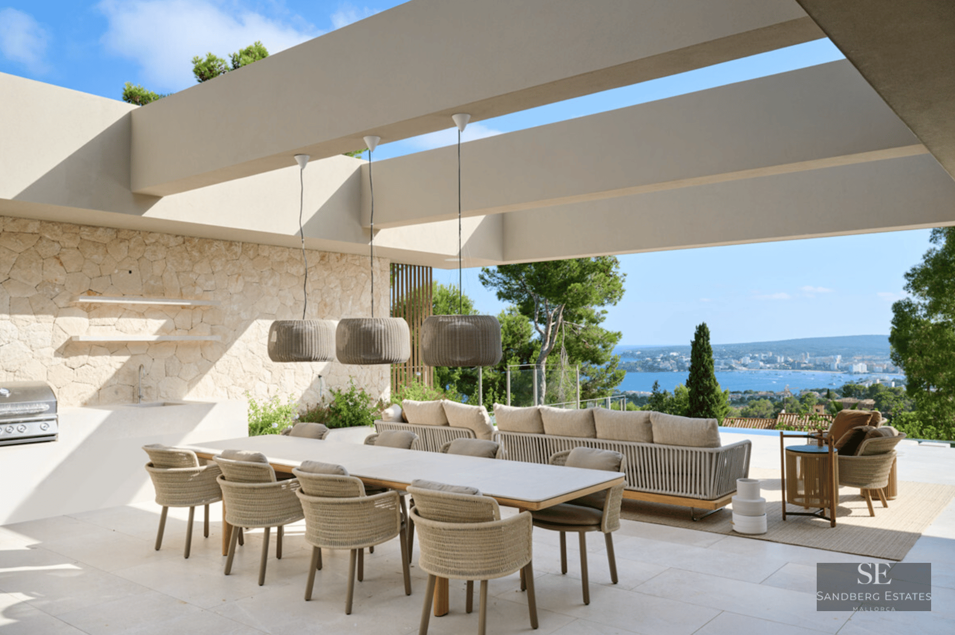 Modern terrace with a long dining table, outdoor kitchen, and sofas overlooking the Mediterranean sea and coastline.