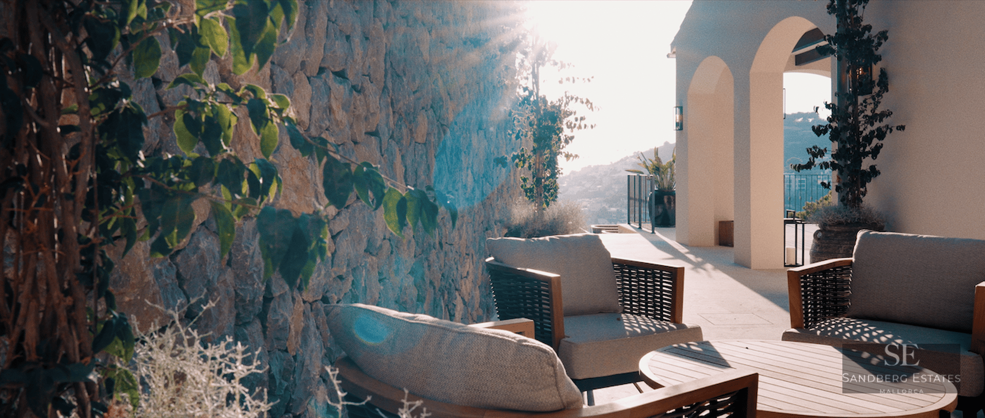 Sun-drenched terrace featuring elegant grey armchairs, a wooden table, a rustic stone wall, and white arched pillars.