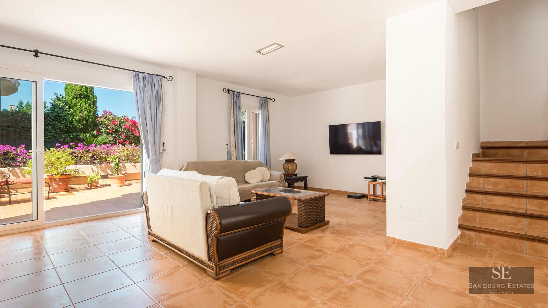 Spacious living room with terracotta floors, sofas, and a large sliding glass door opening to a flowery terrace.
