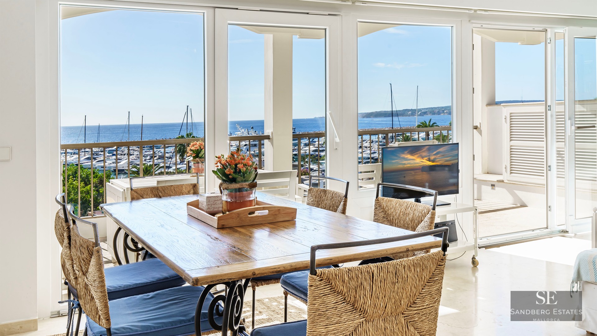 Sunlit dining room with a wooden table and woven chairs looking out over a yacht-filled marina and the blue ocean.