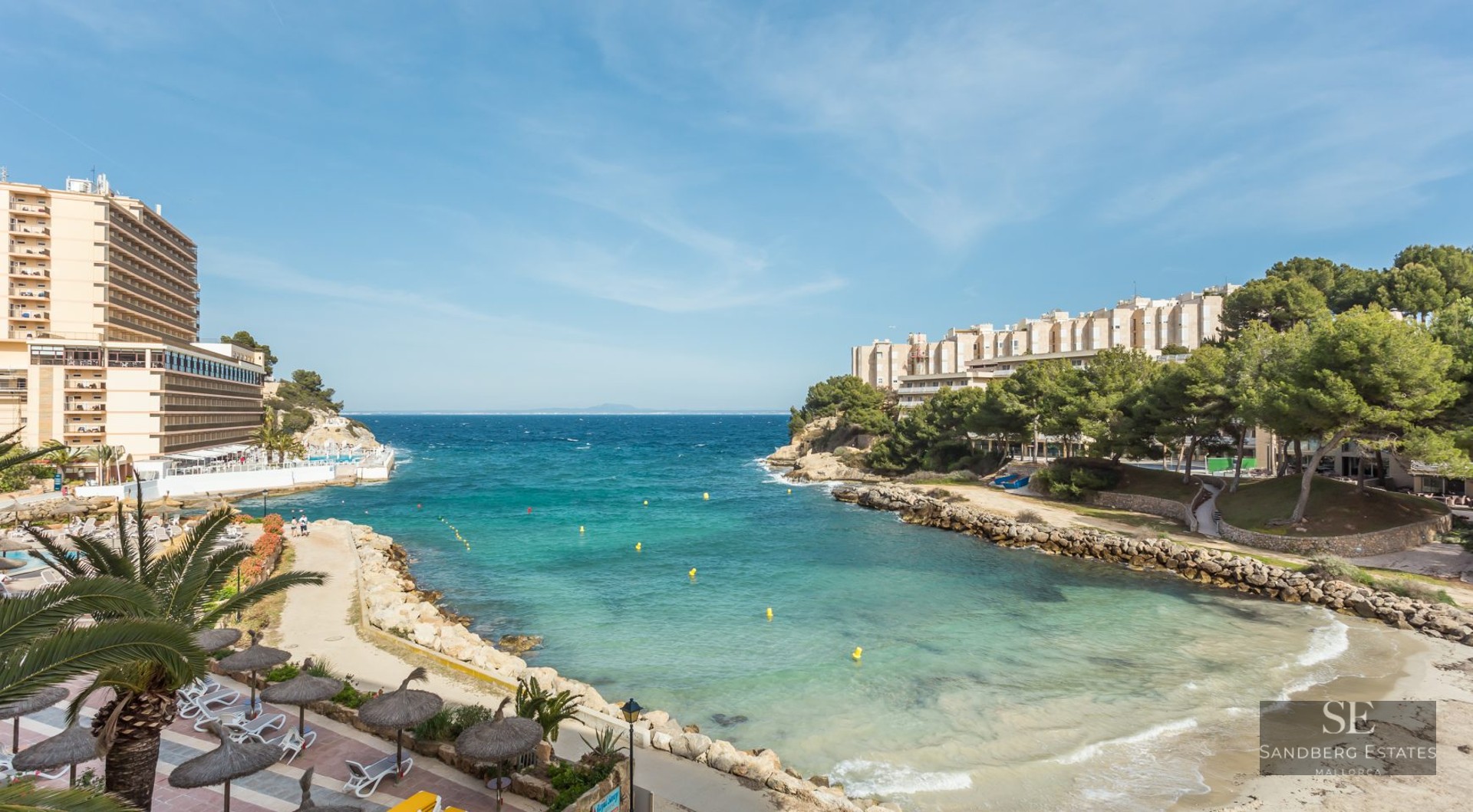 High-angle view of a turquoise sea cove with a small beach, stone walkways, and coastal apartment buildings.