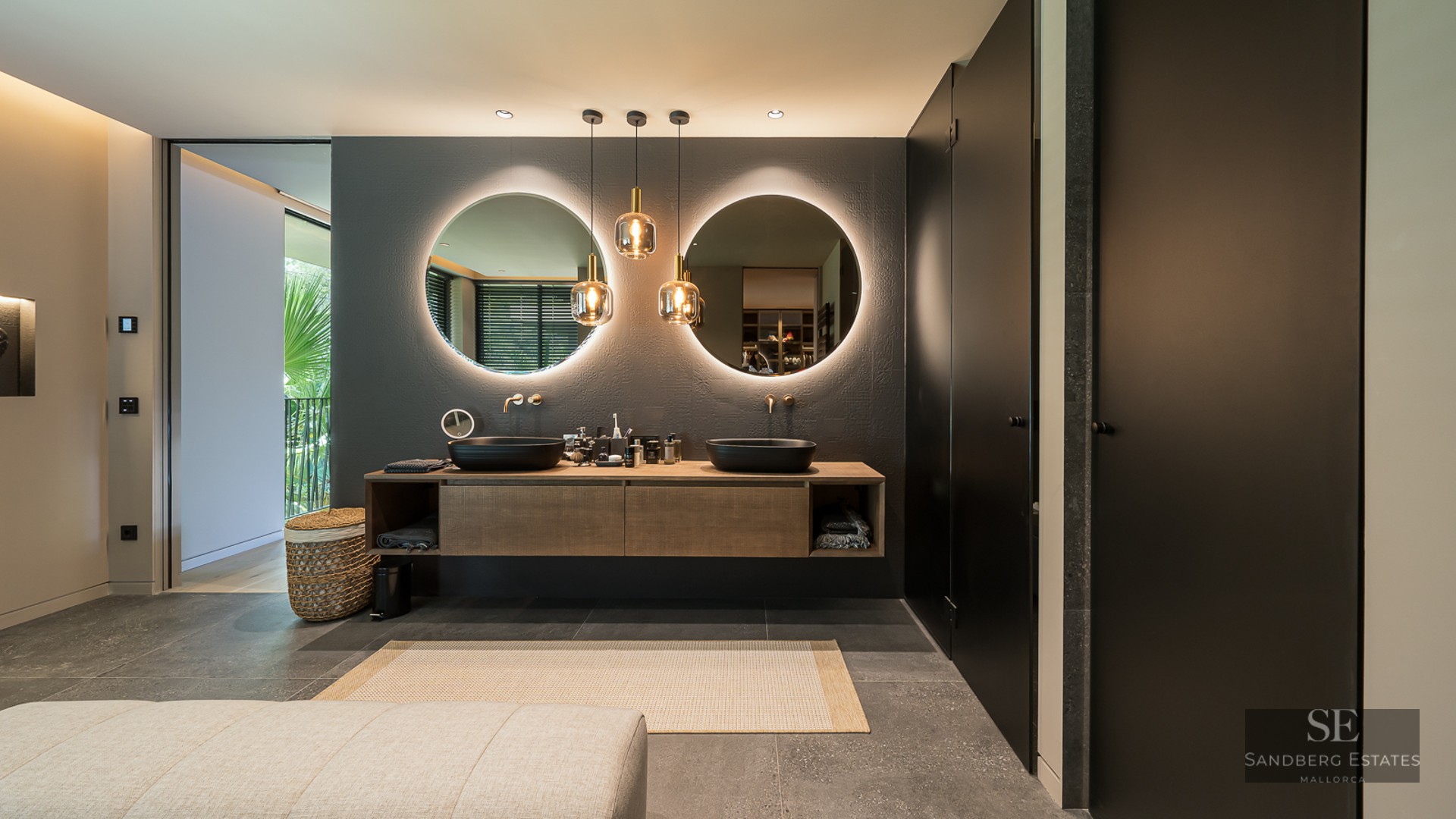 Modern master bathroom featuring two round backlit mirrors, black vessel sinks on a wooden vanity, and grey stone floors.