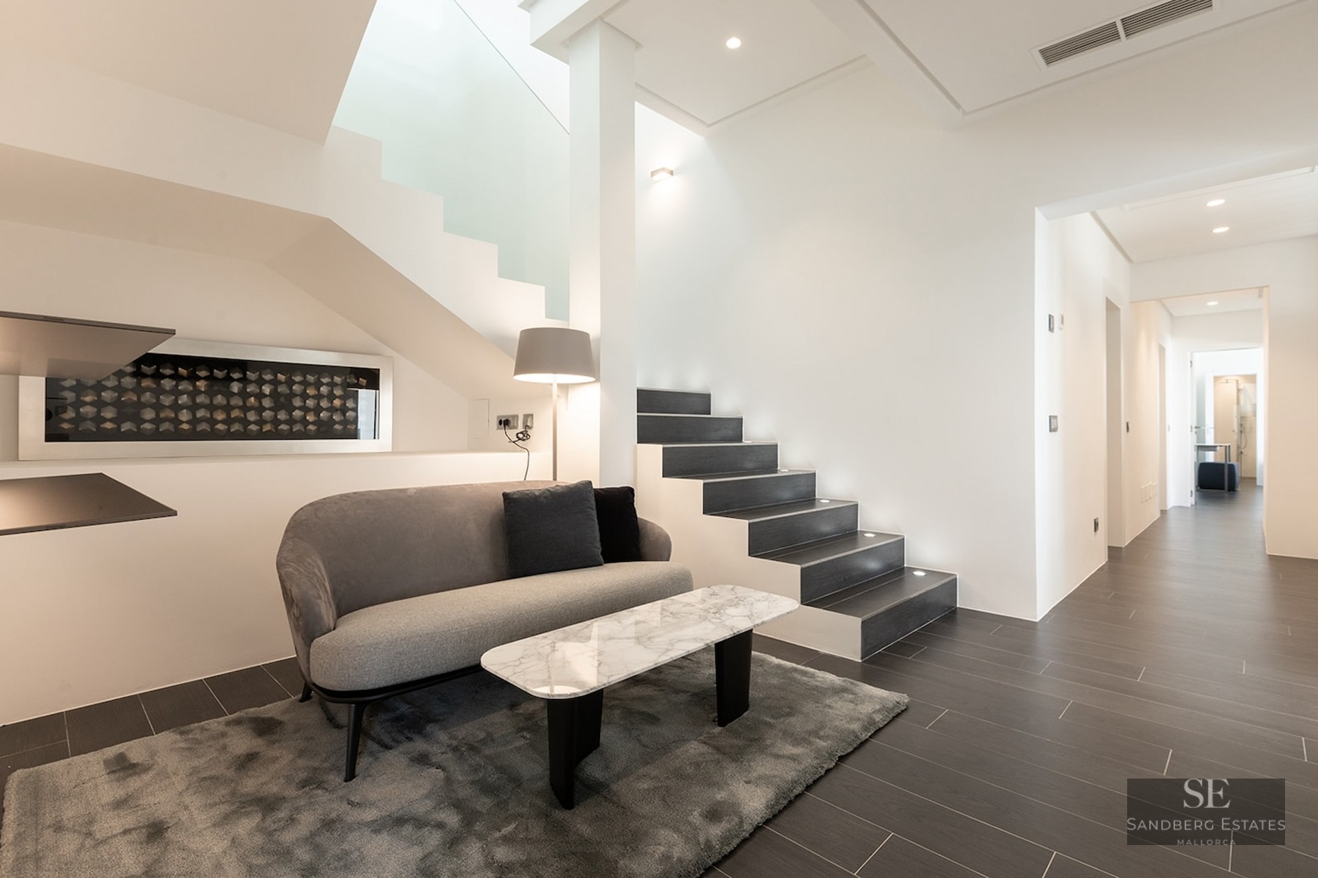 Modern lounge area featuring a grey sofa, marble table, and a contemporary dark-tread staircase with integrated lighting.