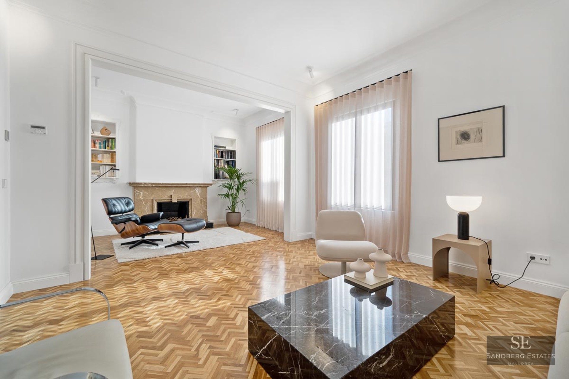Bright living room featuring herringbone wood floors, a black marble coffee table, and an Eames lounge chair by a fireplace.