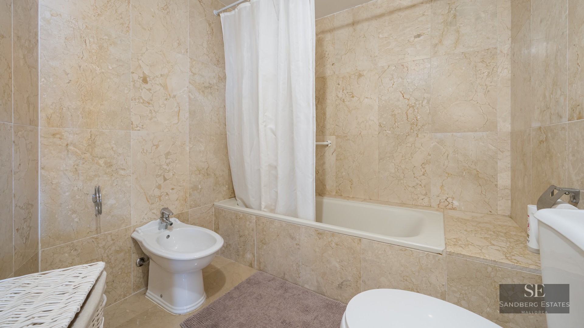 Bathroom featuring beige marble walls, a white bathtub with curtain, and a white ceramic bidet.